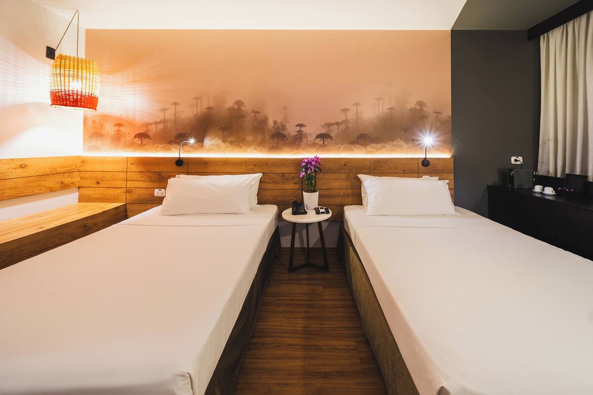 Habitació d'hotel amb dos llits individuals, un capçal de llit de fusta, una taula petita amb un ram de flors i un mural a la paret.