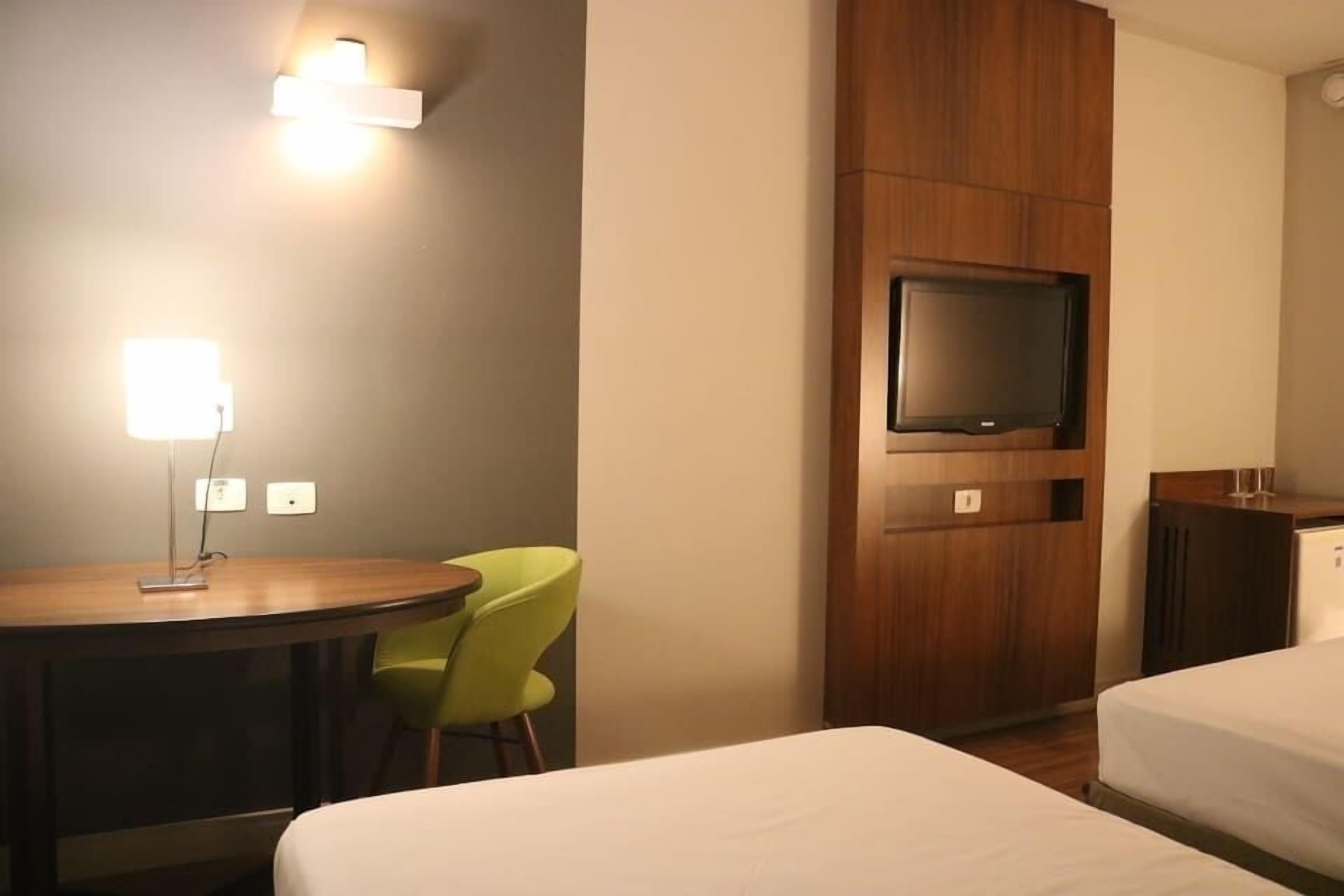 A hotel room with a bed, a desk with a chair, a television mounted on a wooden panel, and a small cabinet.