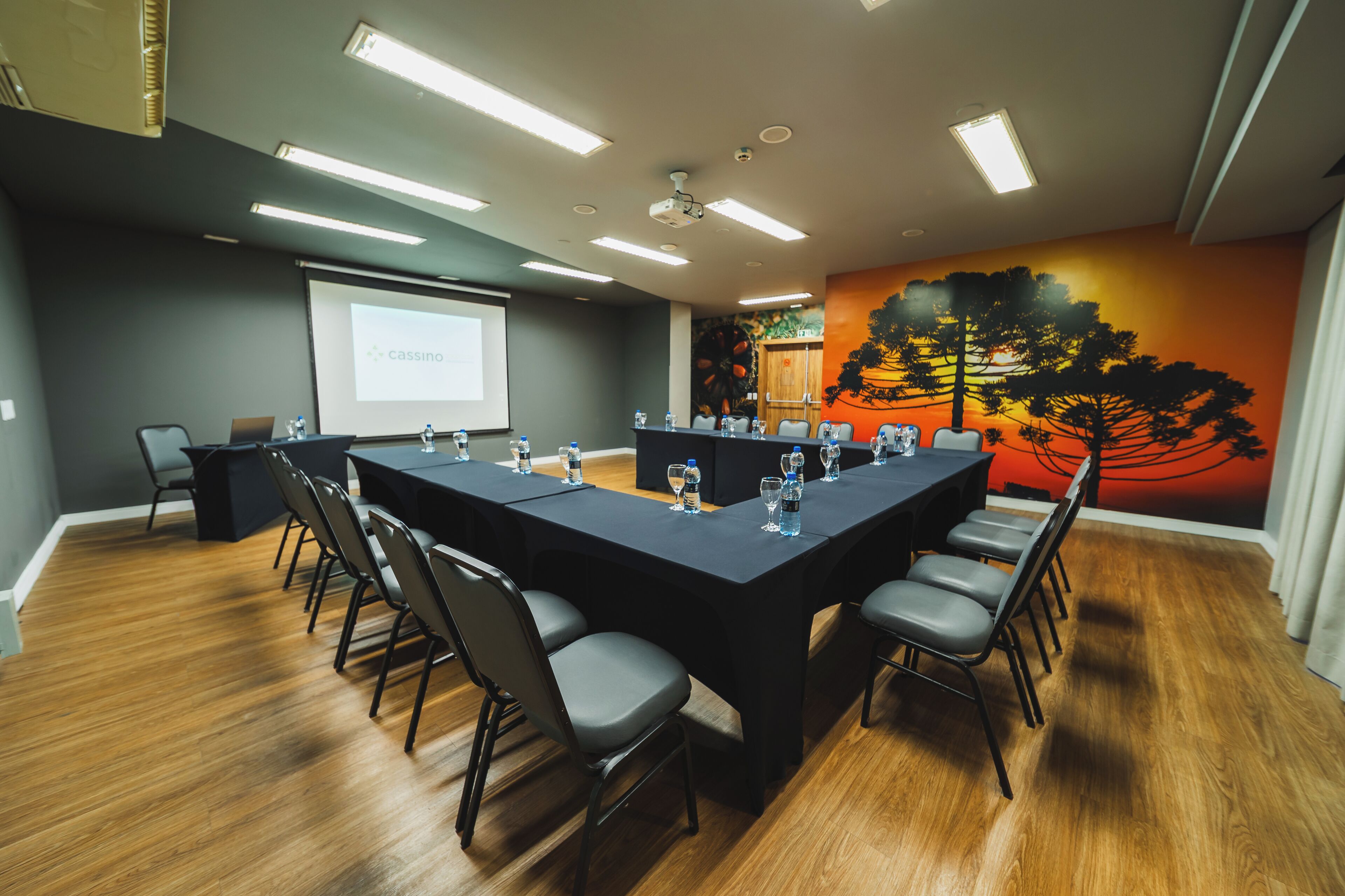 Uma sala de conferências com uma mesa comprida, cadeiras, uma tela de projetor e um mural na parede.