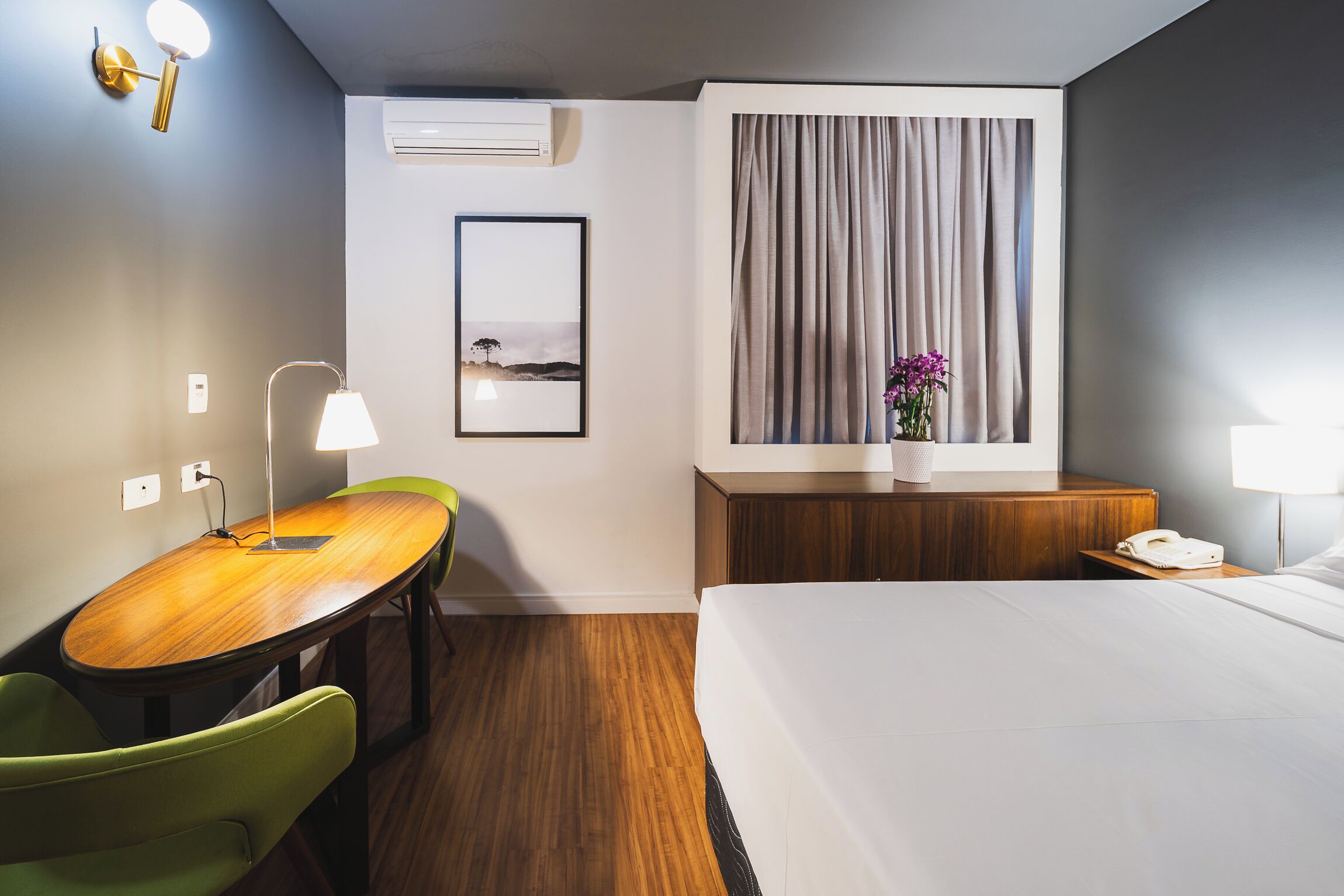 A hotel room with a wooden desk, green chair, bed, bedside table, lamp, and a window with curtains.