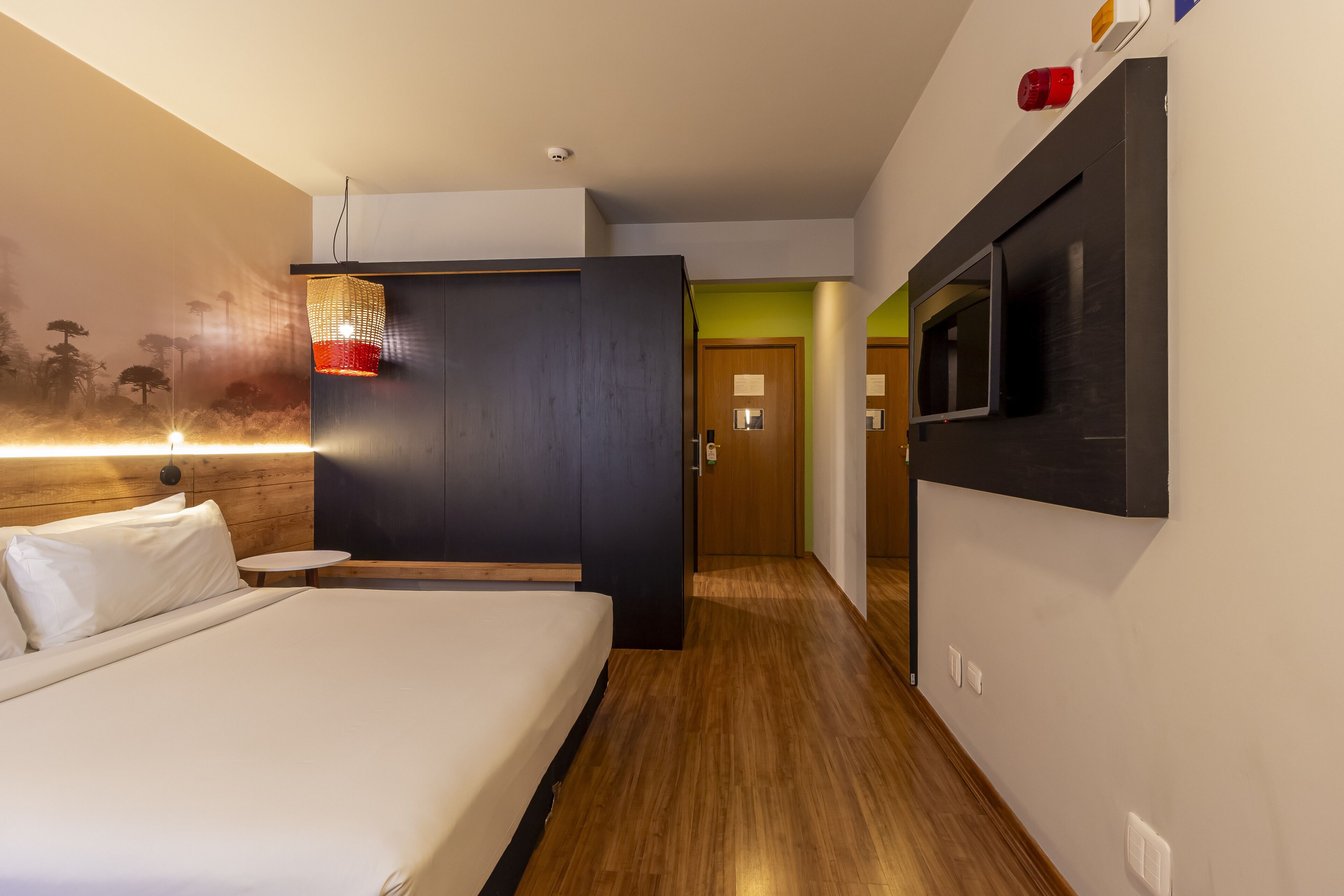 Et moderne hotelværelse med seng, et lille bord, et vægmonteret tv og en dør, der fører til et andet værelse.