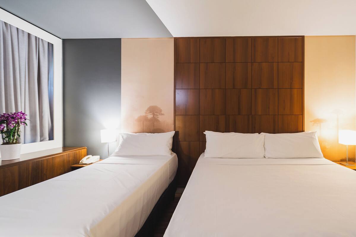 Habitació d'hotel amb dos llits, un panell de fusta al capçal, una petita taula de nit amb una làmpada, i una finestra amb cortines.