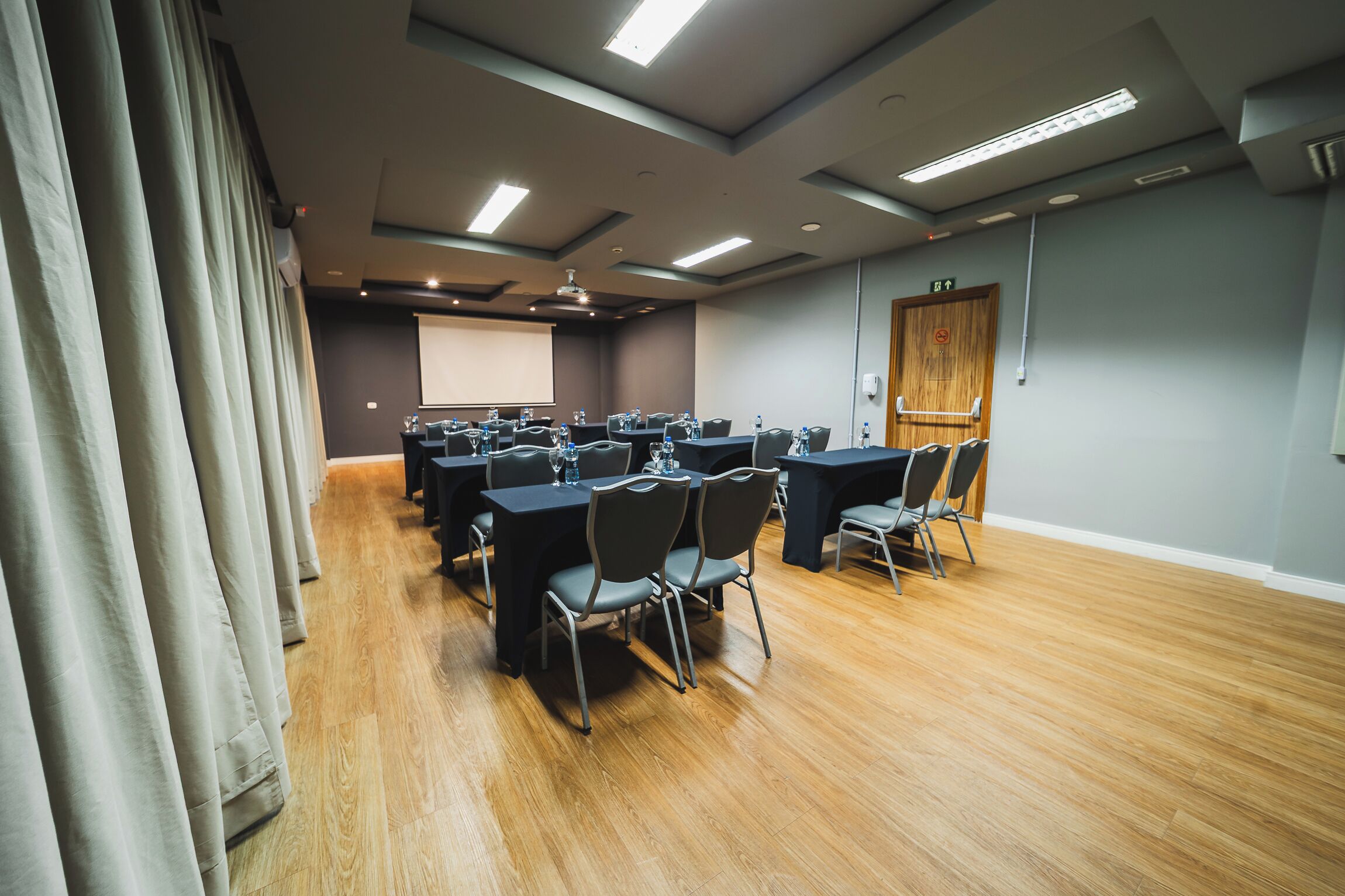 A conference room with tables and chairs, a projector screen, and a wooden floor.