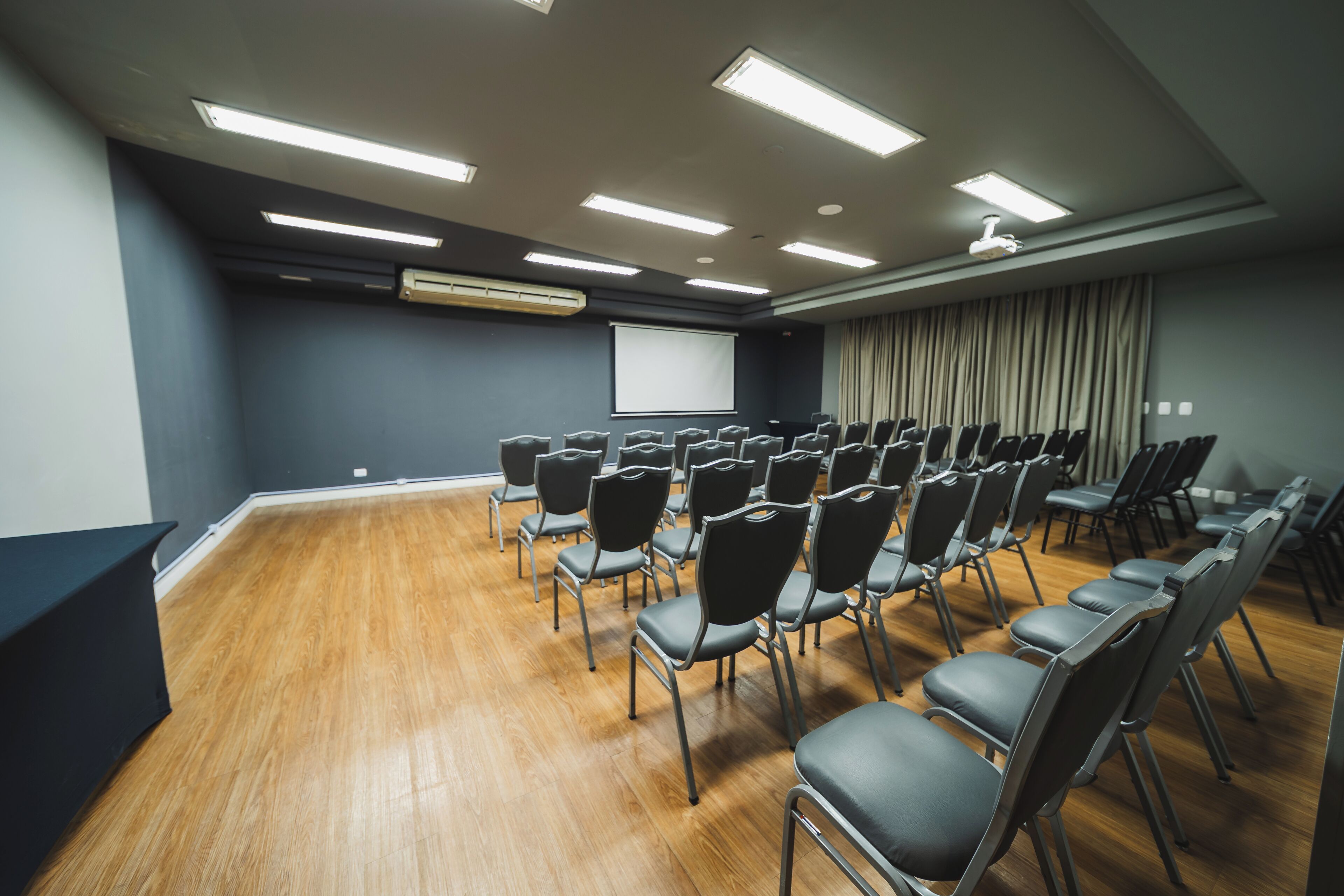 Un salón de conferencias con filas de sillas, una pantalla de proyector y una pantalla grande en la pared.