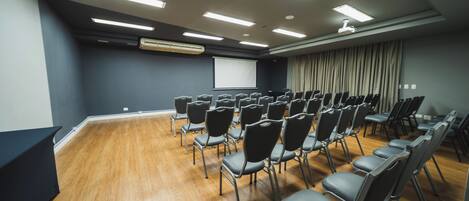 Un salón de conferencias con filas de sillas, una pantalla de proyector y una pantalla grande en la pared.