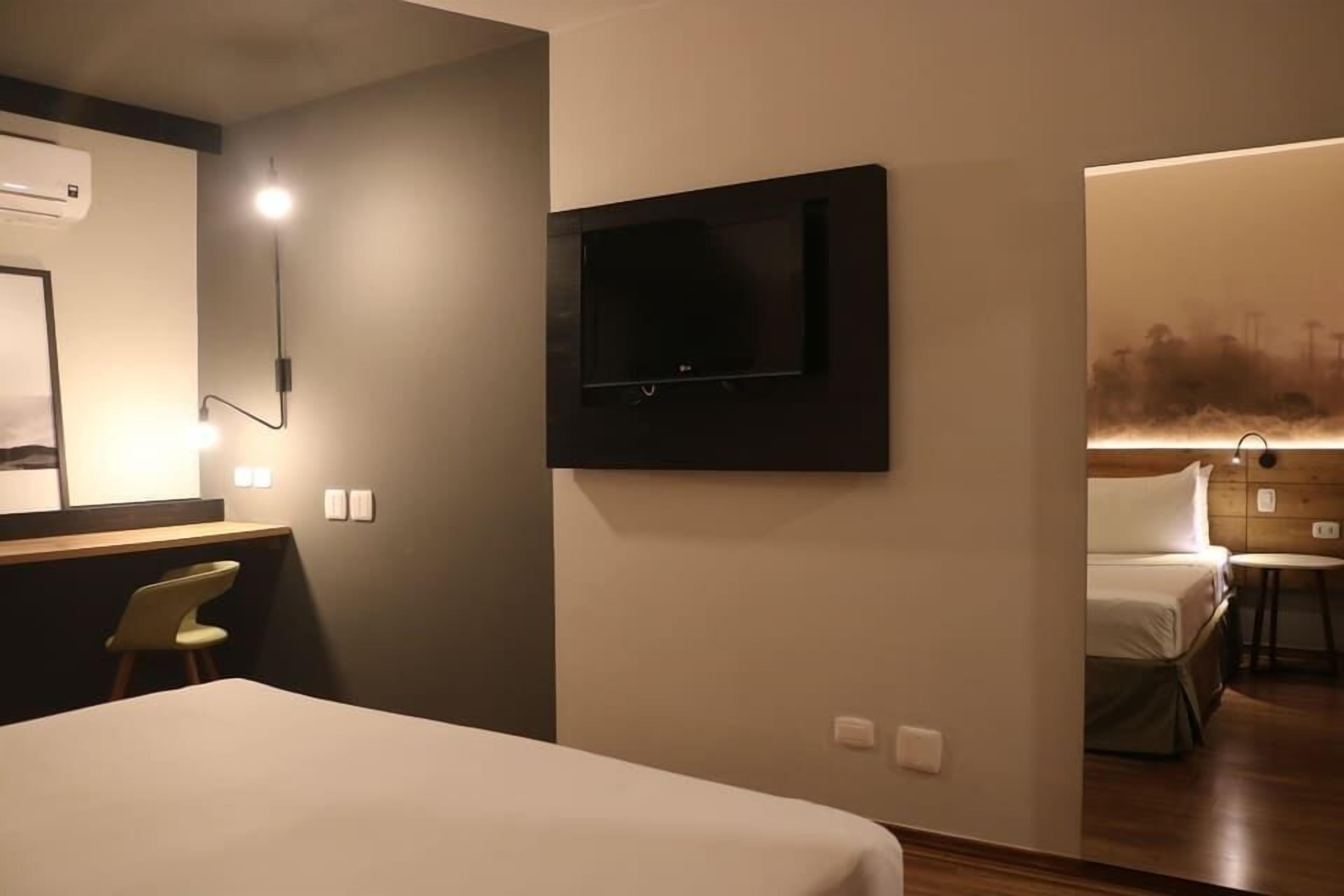 A hotel room with a bed, a desk with a chair, a mounted TV, and a wall-mounted air conditioner.