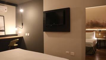 A hotel room with a bed, a desk with a chair, a mounted TV, and a wall-mounted air conditioner.