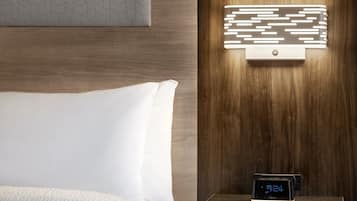 Un lit bien fait, avec une literie blanche, une horloge numérique affichant l’heure et un luminaire mural au design géométrique.