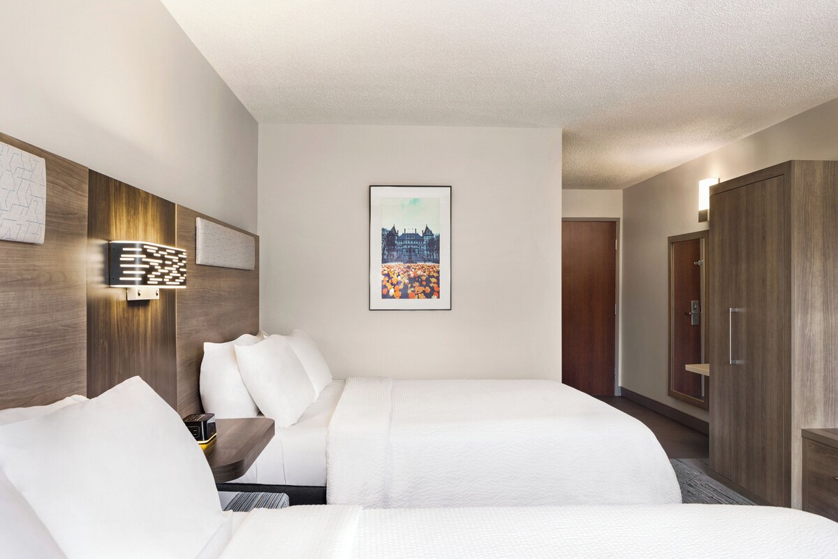 A hotel room with two beds, a wooden headboard, a wall-mounted light, and a framed picture.