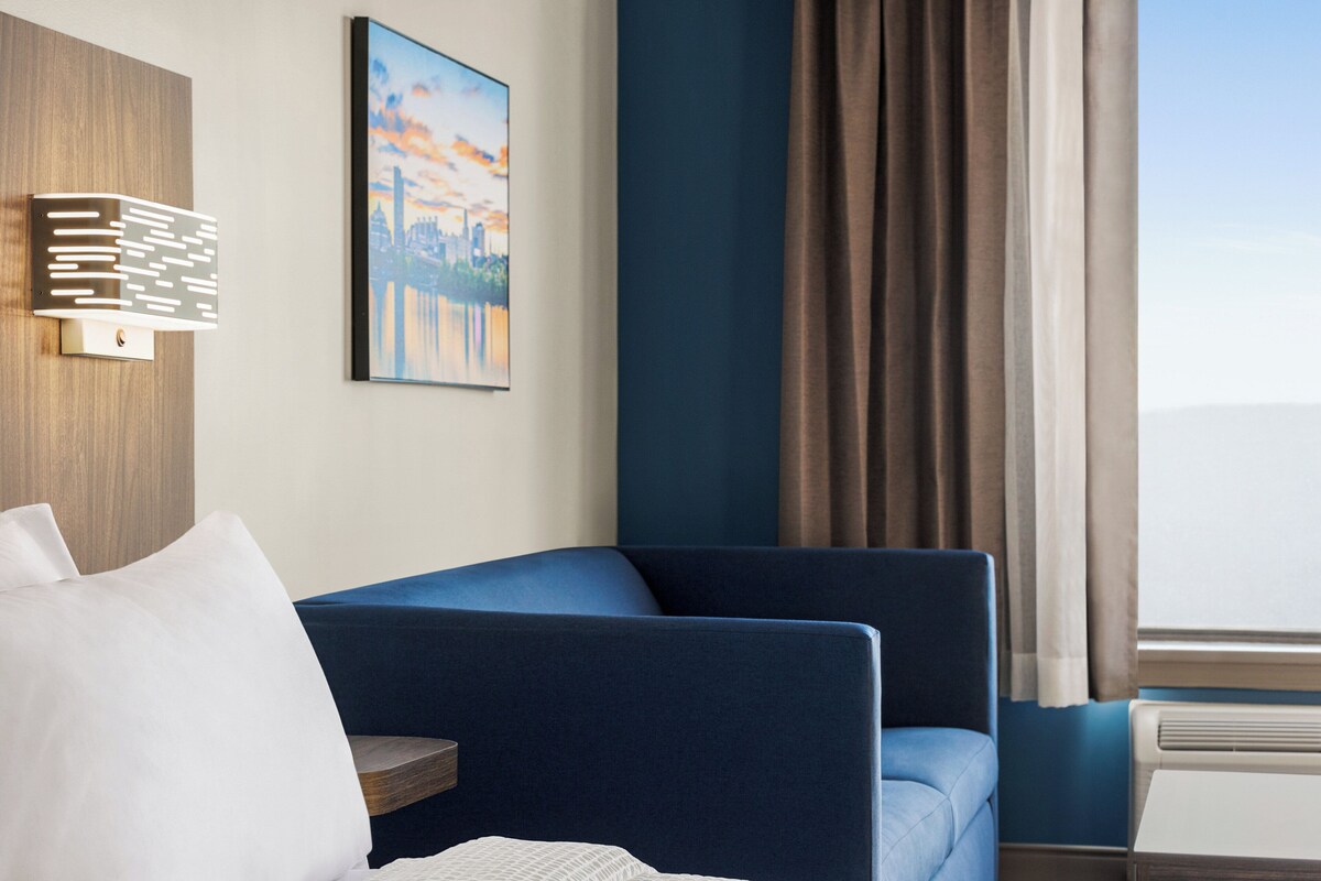 A modern hotel room with a blue sofa, a white pillow, a framed cityscape painting, and a window with curtains.
