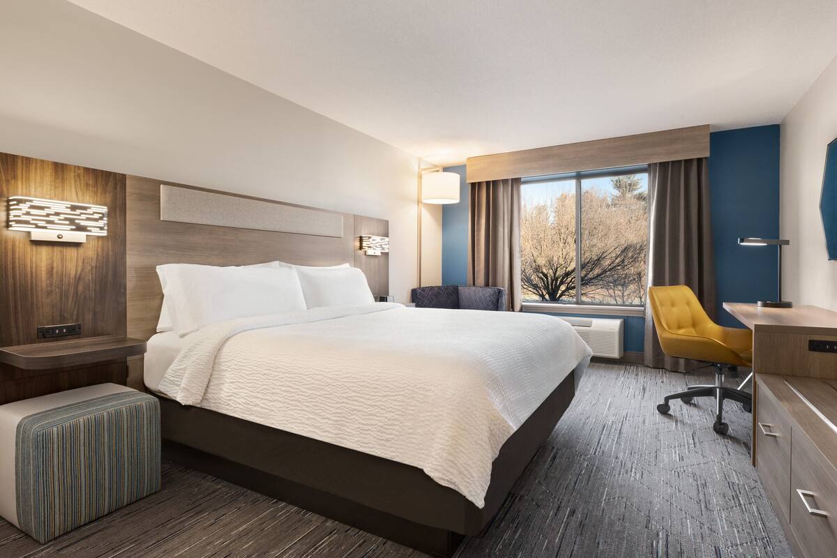 Ein modernes Hotelzimmer mit einem großen Bett, einem Schreibtisch, einem Sessel und Blick auf Bäume durch das Fenster.