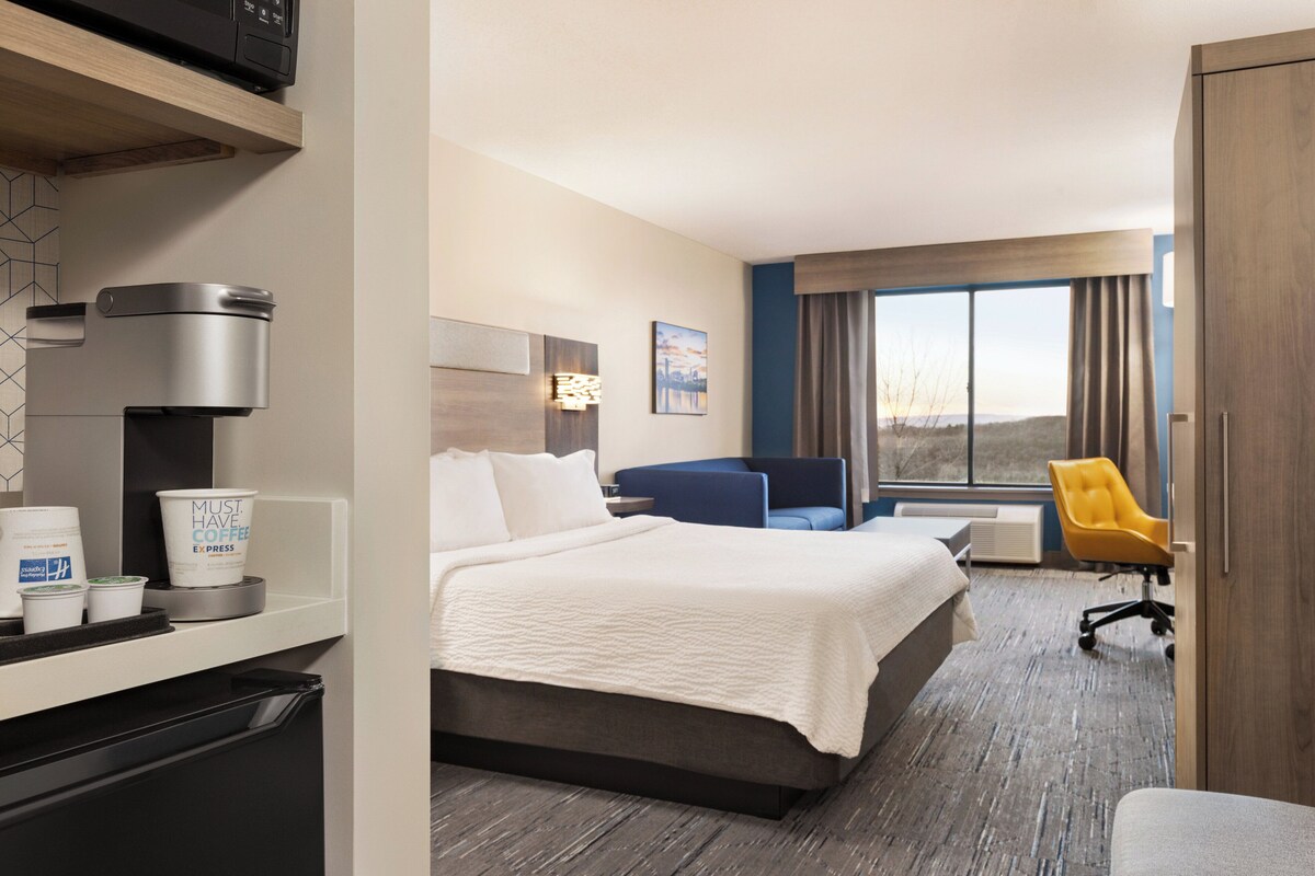 A hotel room with a bed, a desk with a coffee maker, a chair, and a large window with a view.
