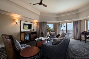 A hotel room with a large sofa, a television, a dining table, and a balcony with a view of the ocean.