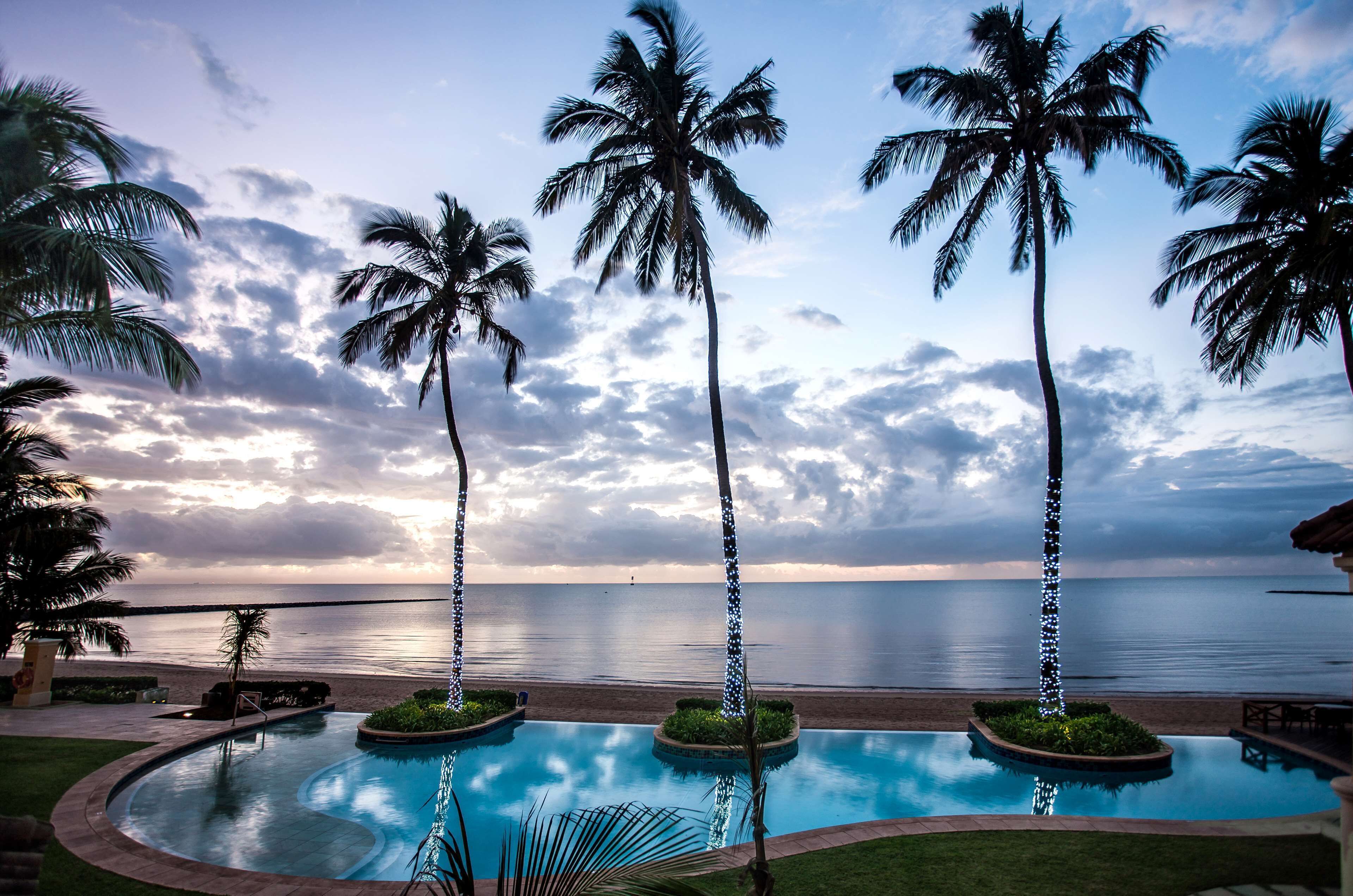 A pool with palm trees and a view of the ocean at sunset.