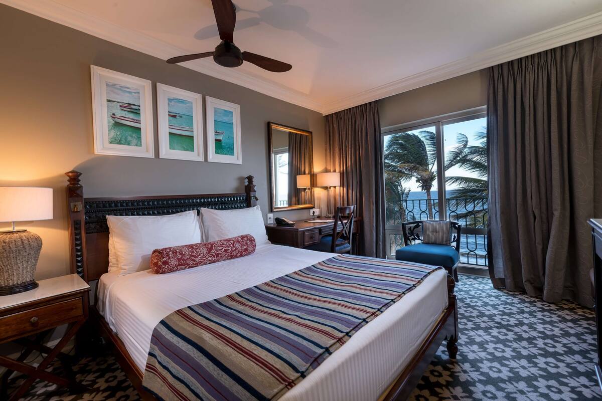 A hotel room with a large bed, a desk, a chair, and a balcony with a view of palm trees.