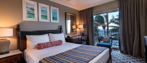 A hotel room with a large bed, a desk, a chair, and a balcony with a view of palm trees.