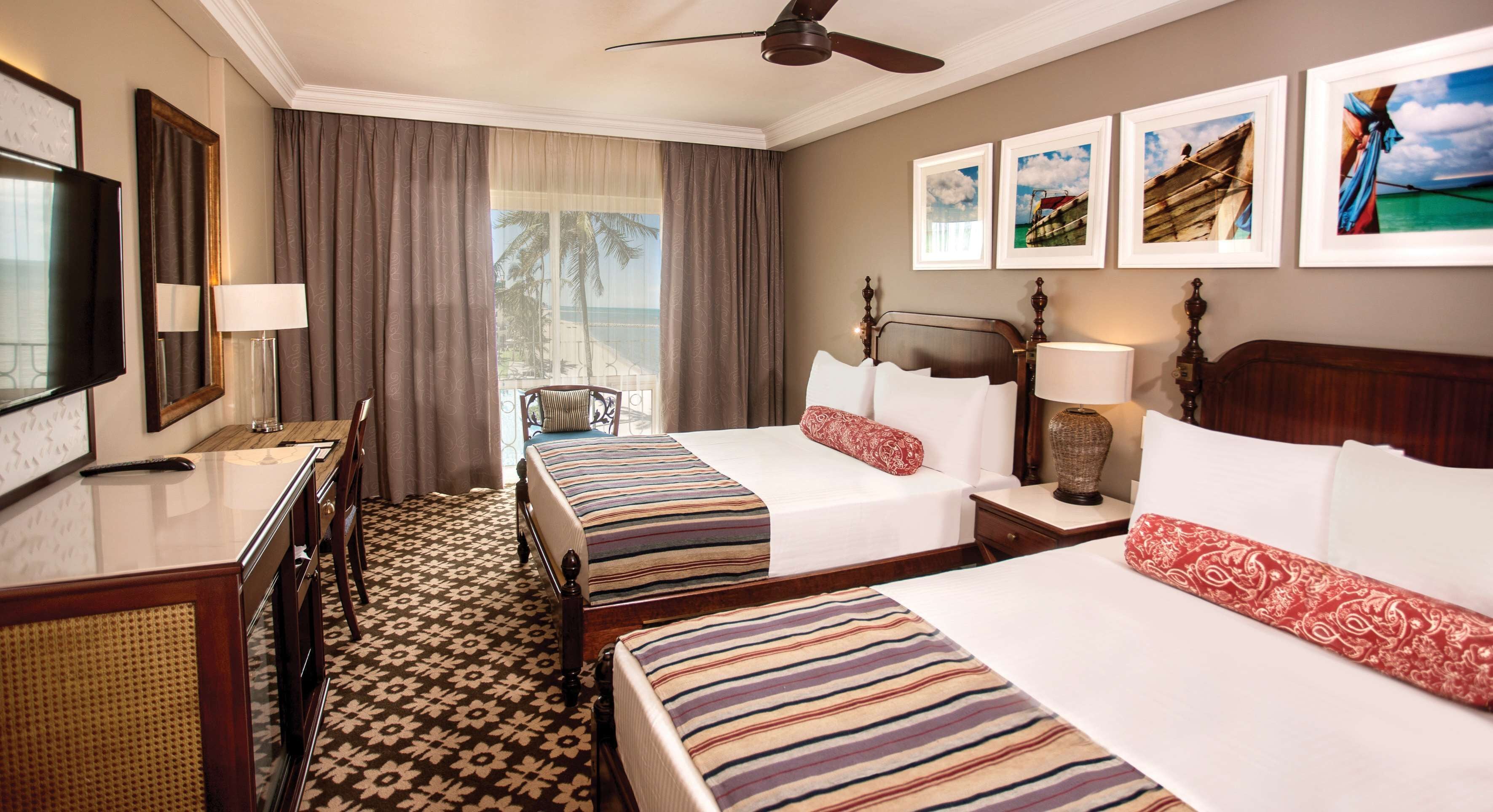 A hotel room with two beds, a TV, a desk, a chair, and a balcony with a view of the ocean.