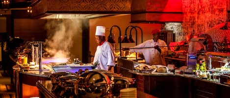Una cocina profesional con chefs trabajando, equipada con electrodomésticos de acero inoxidable y una variedad de alimentos a la vista.