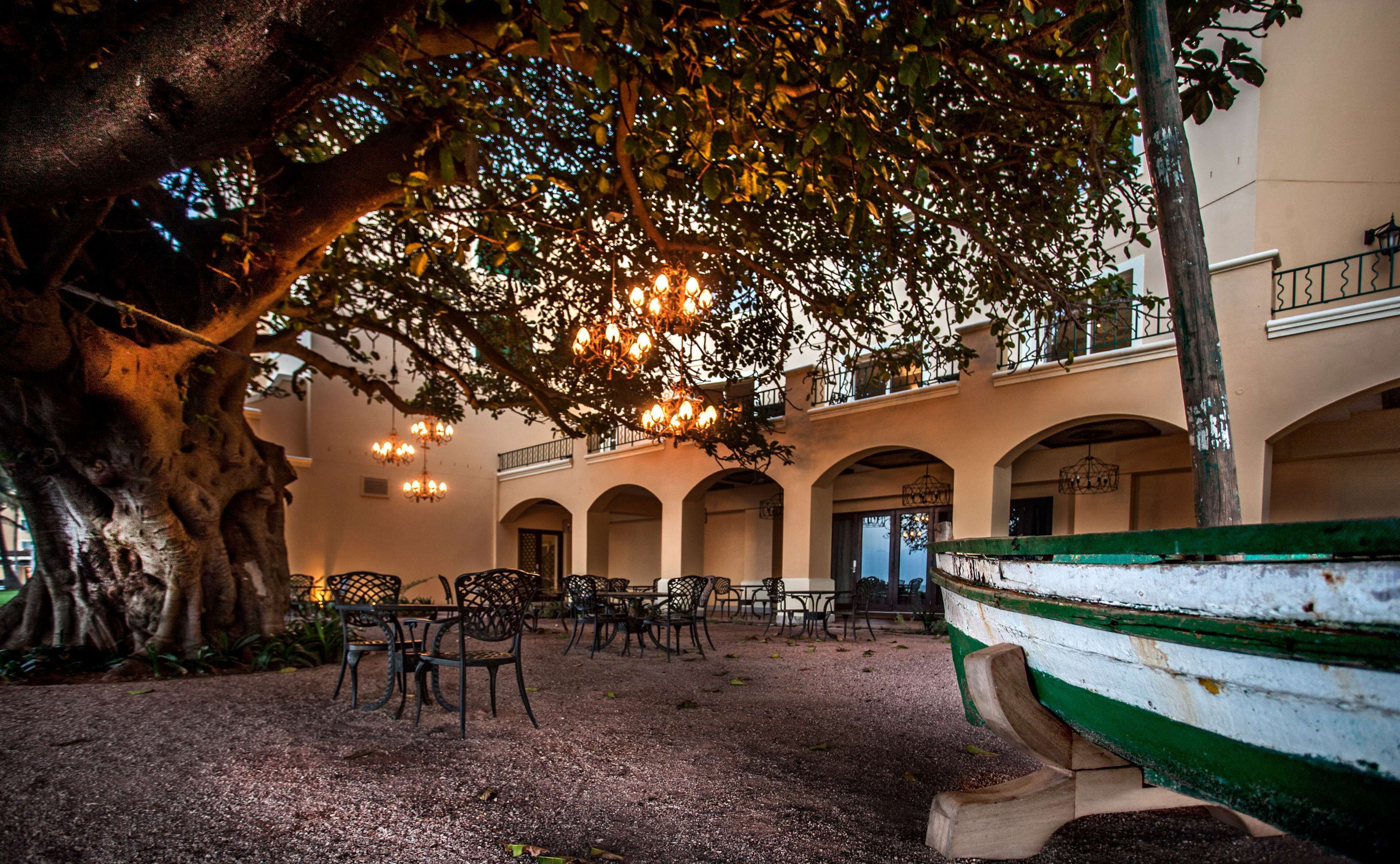 An outdoor seating area with tables and chairs under a tree, surrounded by a building with arches and a boat on the right.