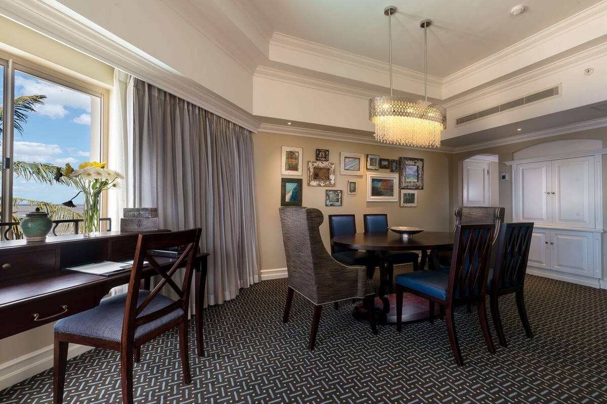 A dining area with a round table, chairs, and a chandelier. There are framed pictures on the wall and a window with curtains.