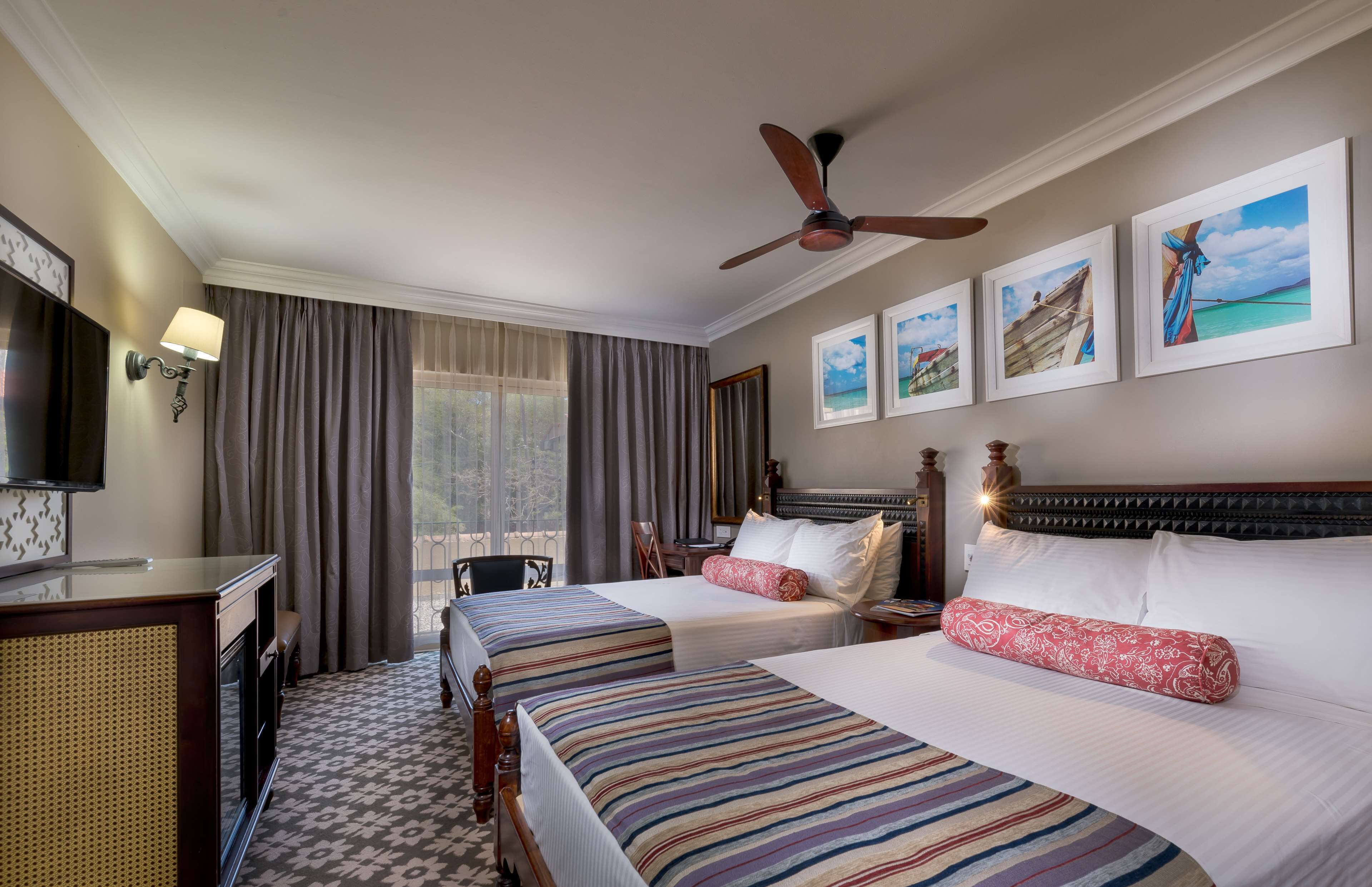 A hotel room with two beds, a desk, a television, and a ceiling fan.