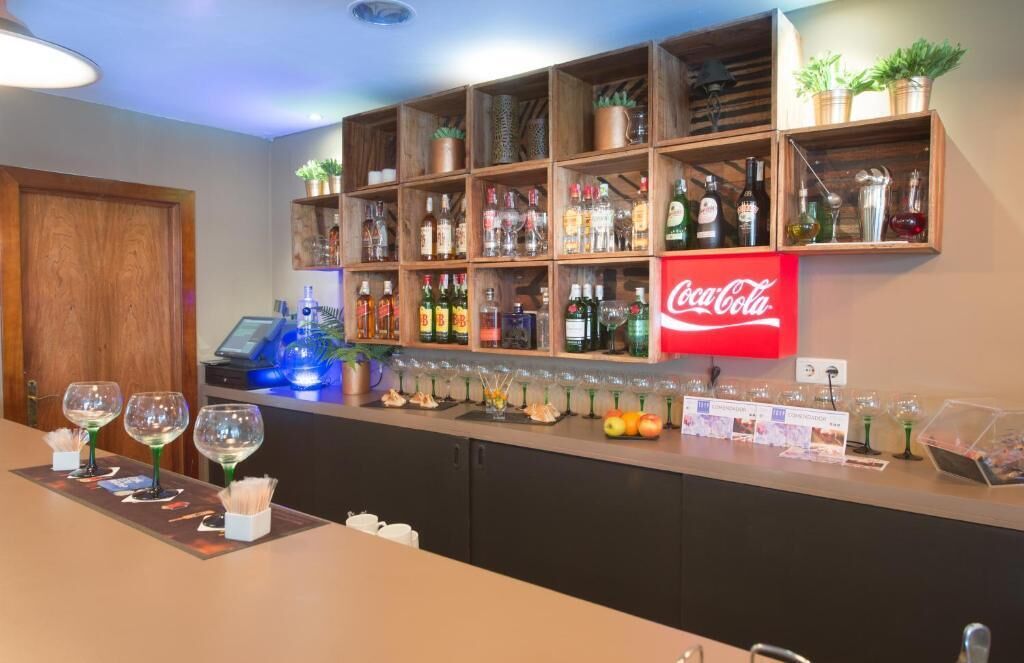 En bar med hylder fyldt med flasker spiritus, et Coca-Cola skilt og en bardisk med vinglas og frugt.