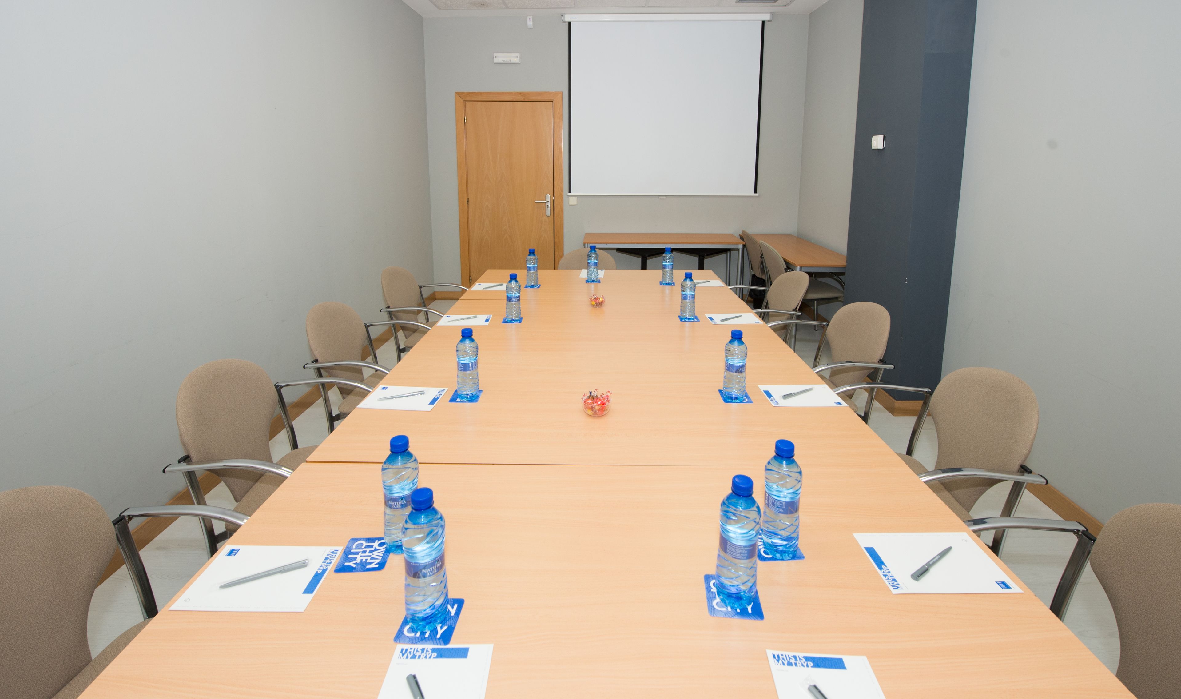 A conference room with a long table, chairs, water bottles, and a projector screen.