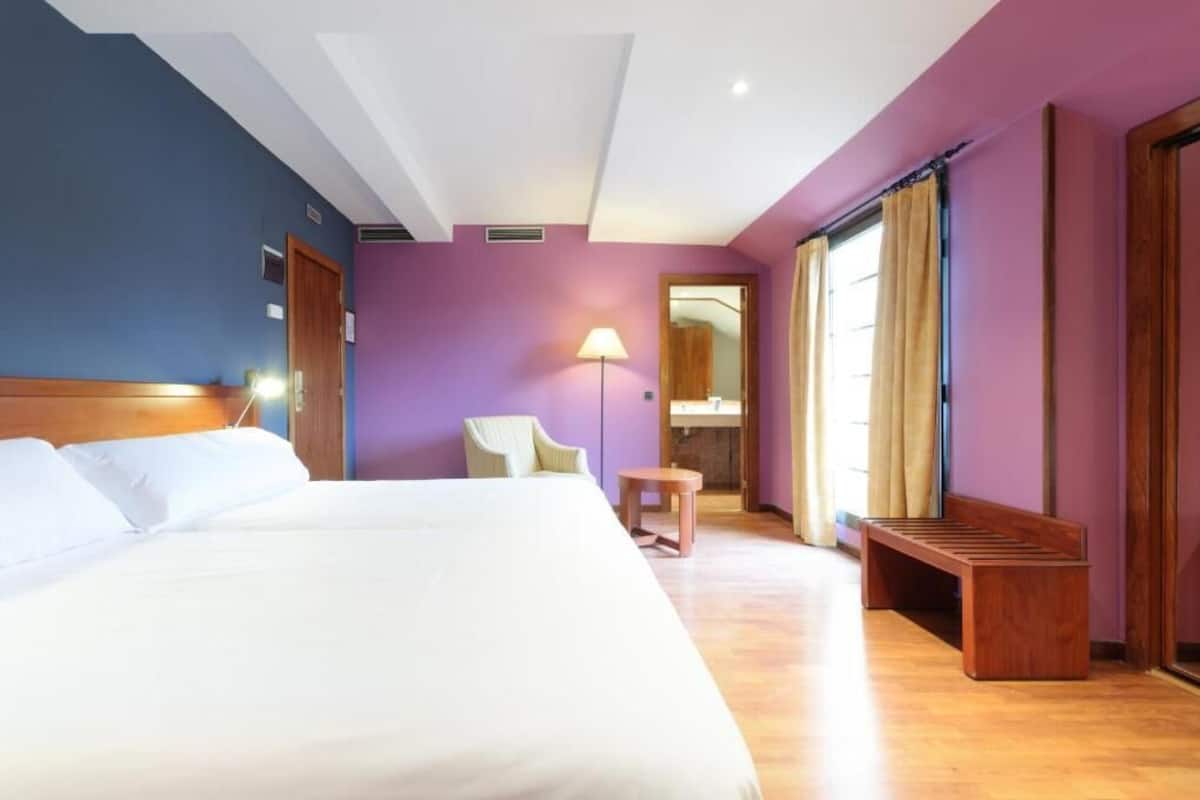 A hotel room with a large bed, a chair, a small table, and a bench.