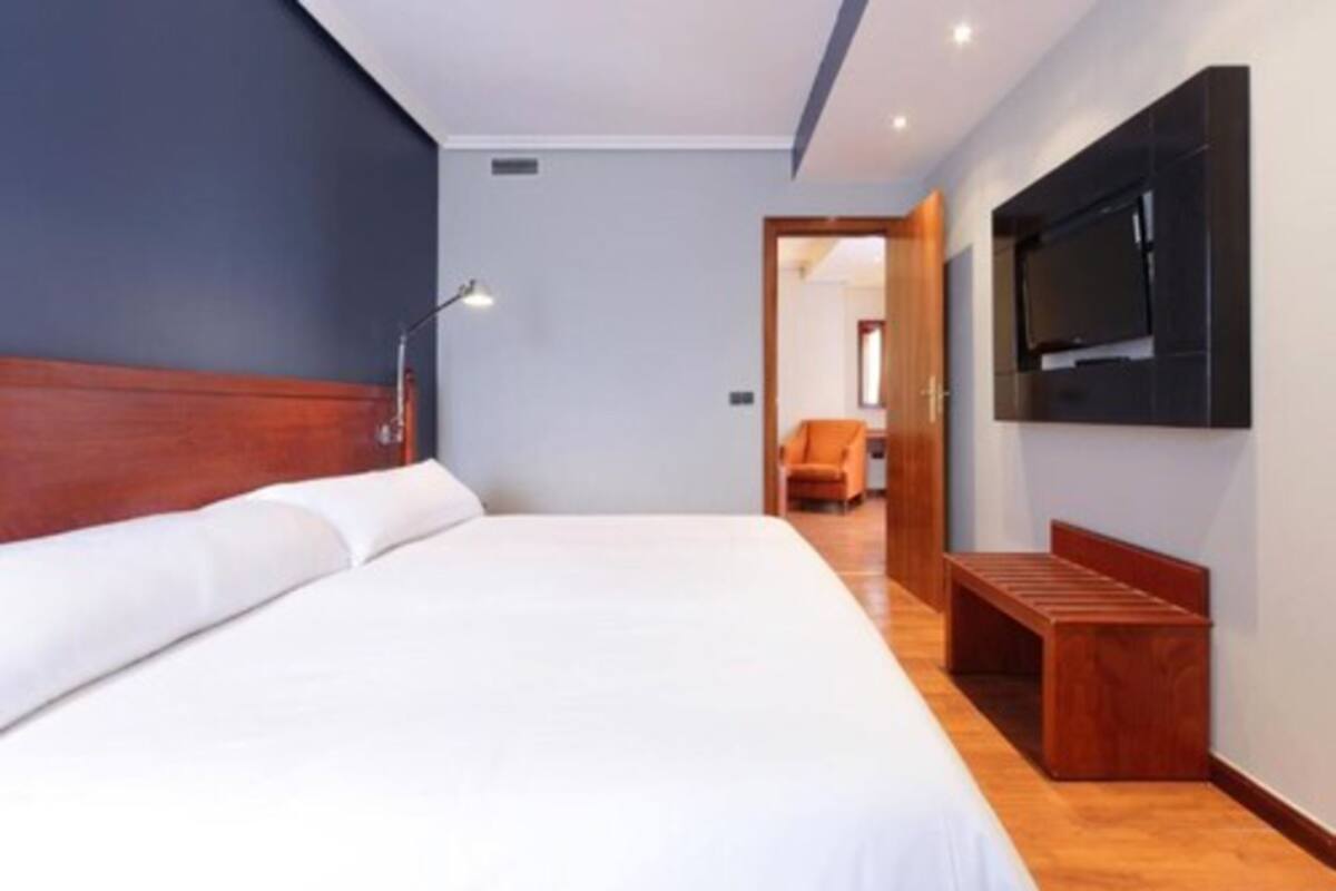 A modern hotel room with a large bed, a wooden bench, a flat-screen TV mounted on the wall, and a door leading to another room.