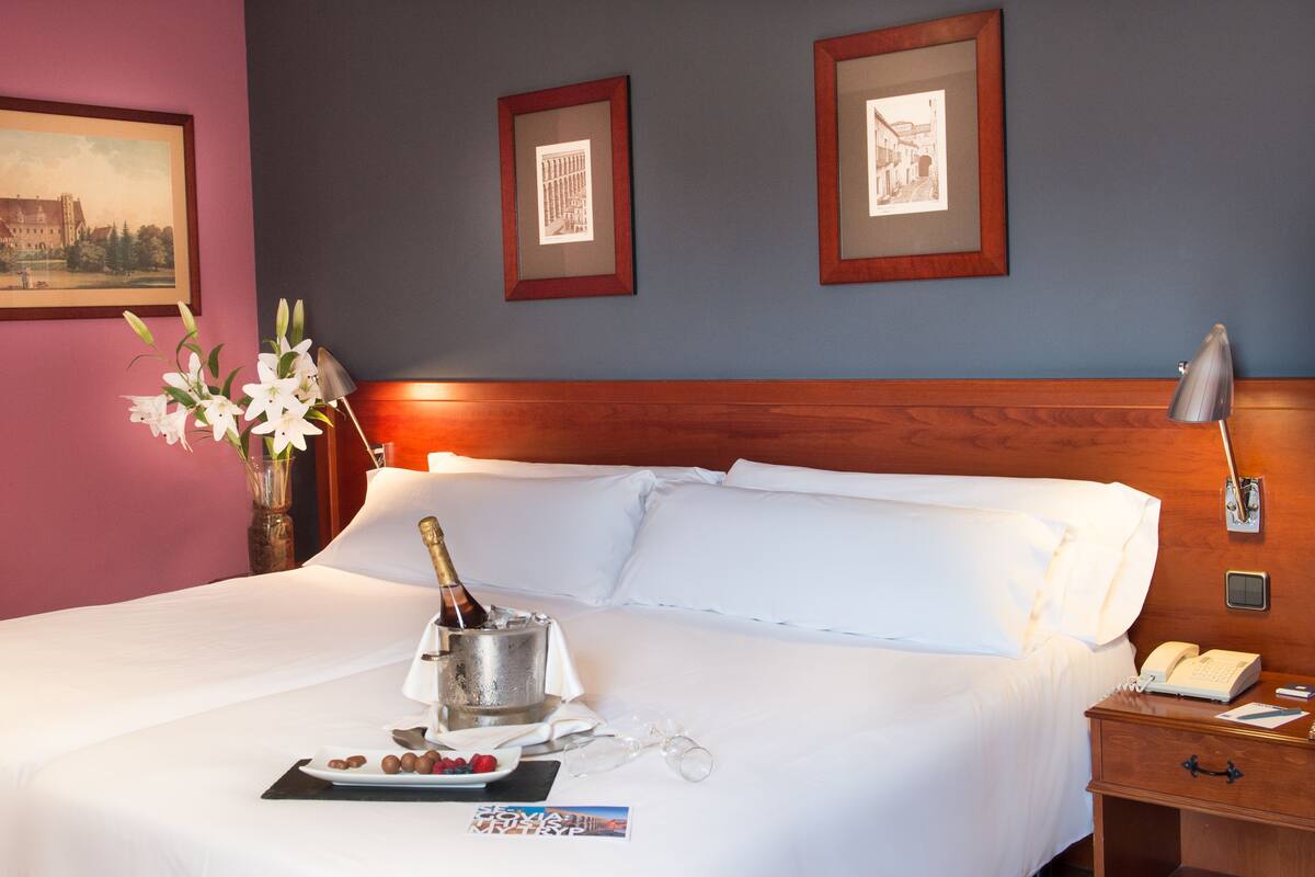 A hotel room with a bed, a nightstand, a lamp, a vase with flowers, a bottle of champagne, and framed pictures on the wall.