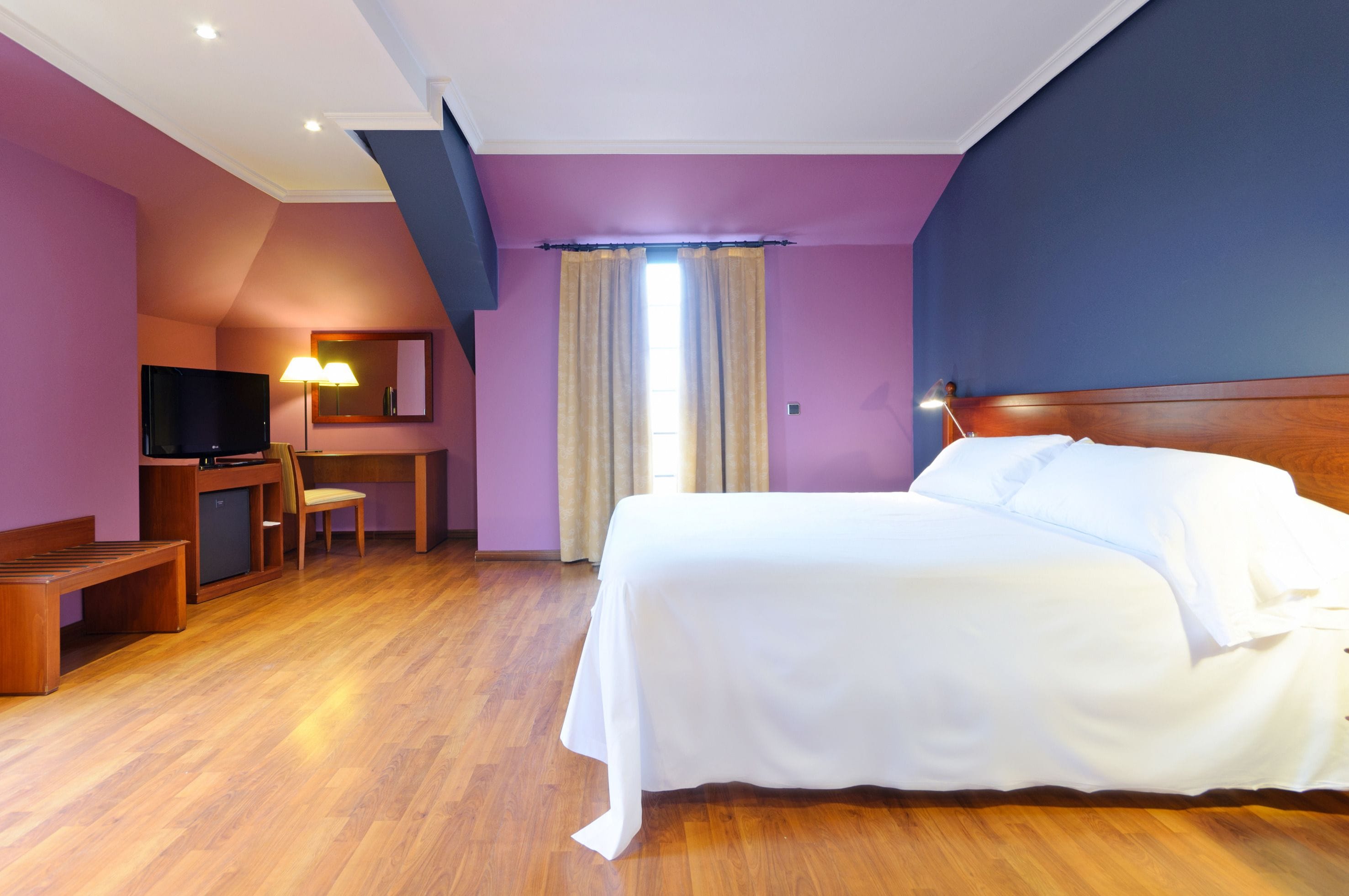 A hotel room with a bed, desk, chair, TV, and curtains.