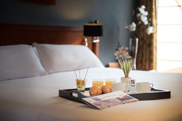 Una cama bien hecha con sábanas blancas, una bandeja con cruasanes, jugo y una taza, y un florero con flores en una habitación de hotel.
