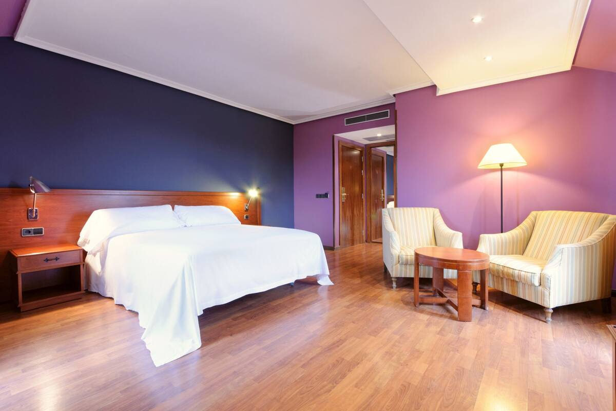 A hotel room with a bed, nightstand, lamp, and two chairs.