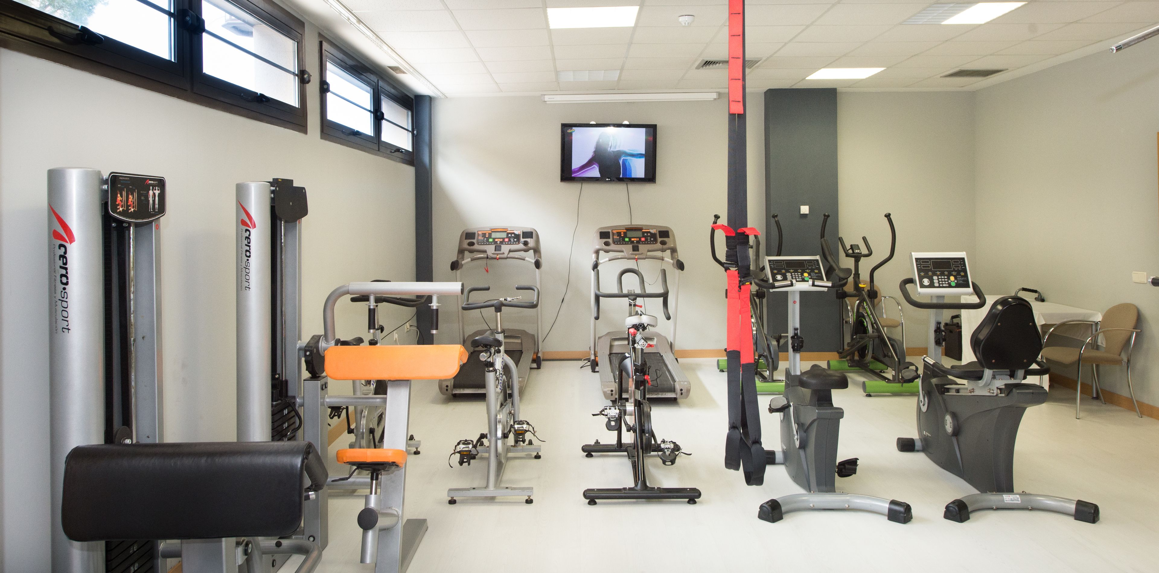 A gym with various exercise equipment, a television on the wall, and a window providing natural light.