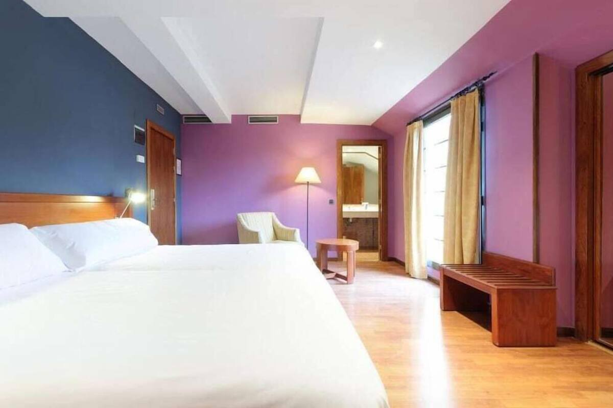 A hotel room with a large bed, a chair, a small table, and a bench.