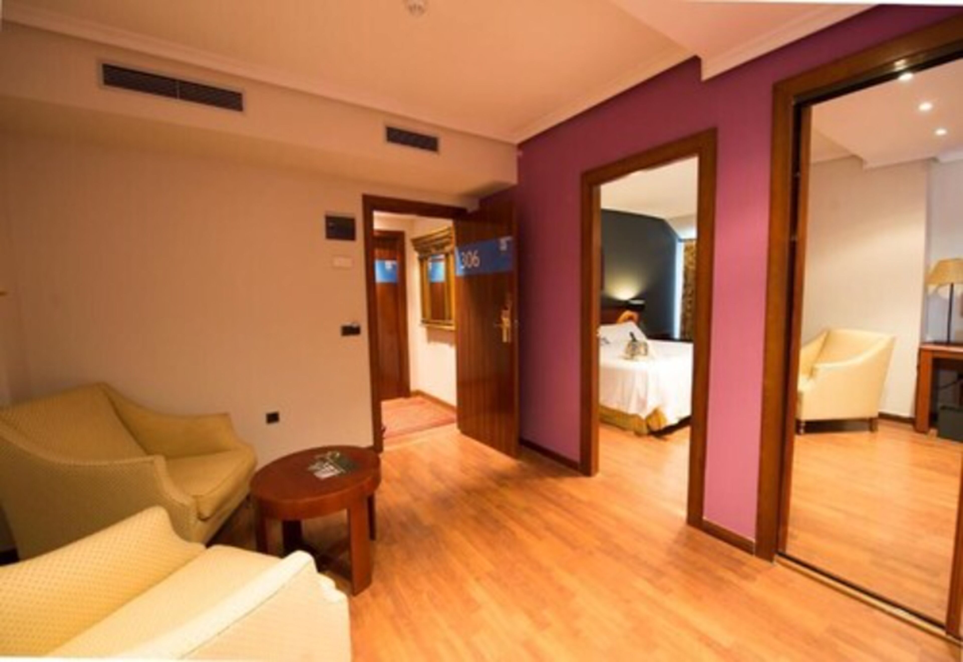 Habitació d'hotel amb sòl de fusta, un llit, una cadira, una taula petita i una porta que dona a una altra habitació.