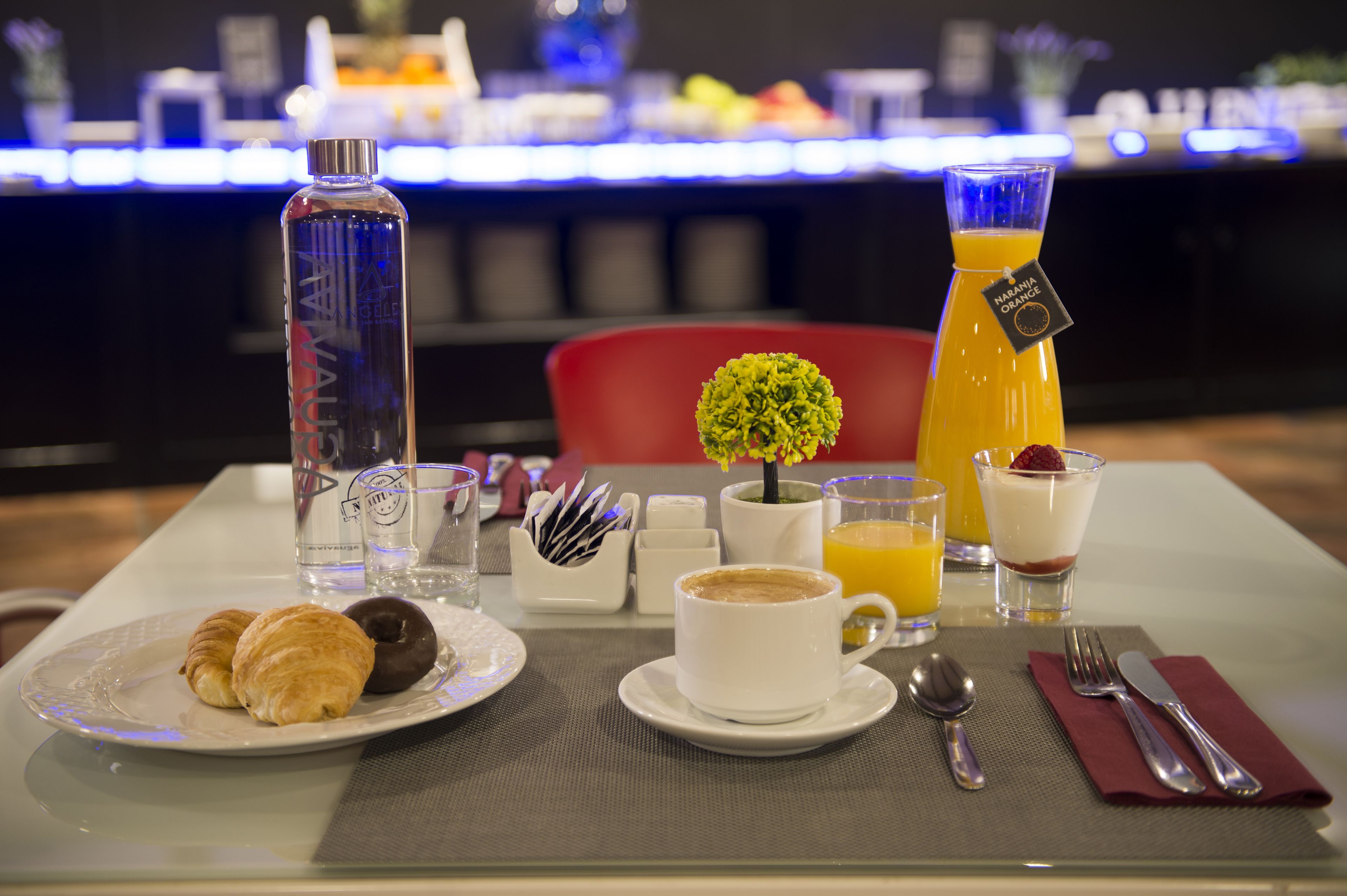 A breakfast table with croissants, a cup of coffee, a glass of orange juice, a bottle of water, and a small plant.