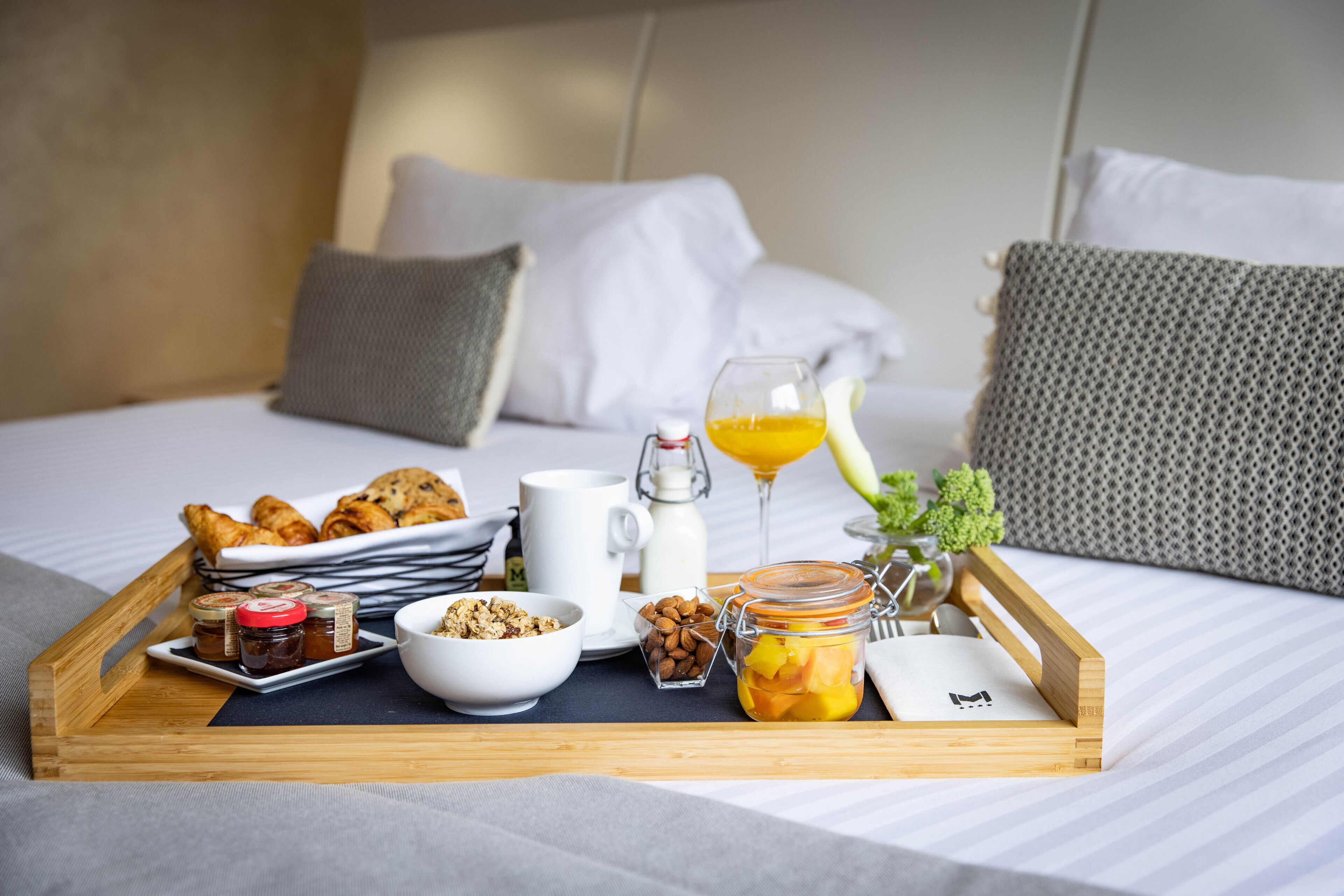 Un plateau de petit-déjeuner avec des viennoiseries, un verre de jus d’orange, un bol de céréales, un pot de confiture et une petite assiette d’amandes, le tout posé sur un lit avec une literie blanche et des oreillers gris.