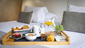 Un plateau de petit-déjeuner avec des viennoiseries, un verre de jus d’orange, un bol de céréales, un pot de confiture et une petite assiette d’amandes, le tout posé sur un lit avec une literie blanche et des oreillers gris.