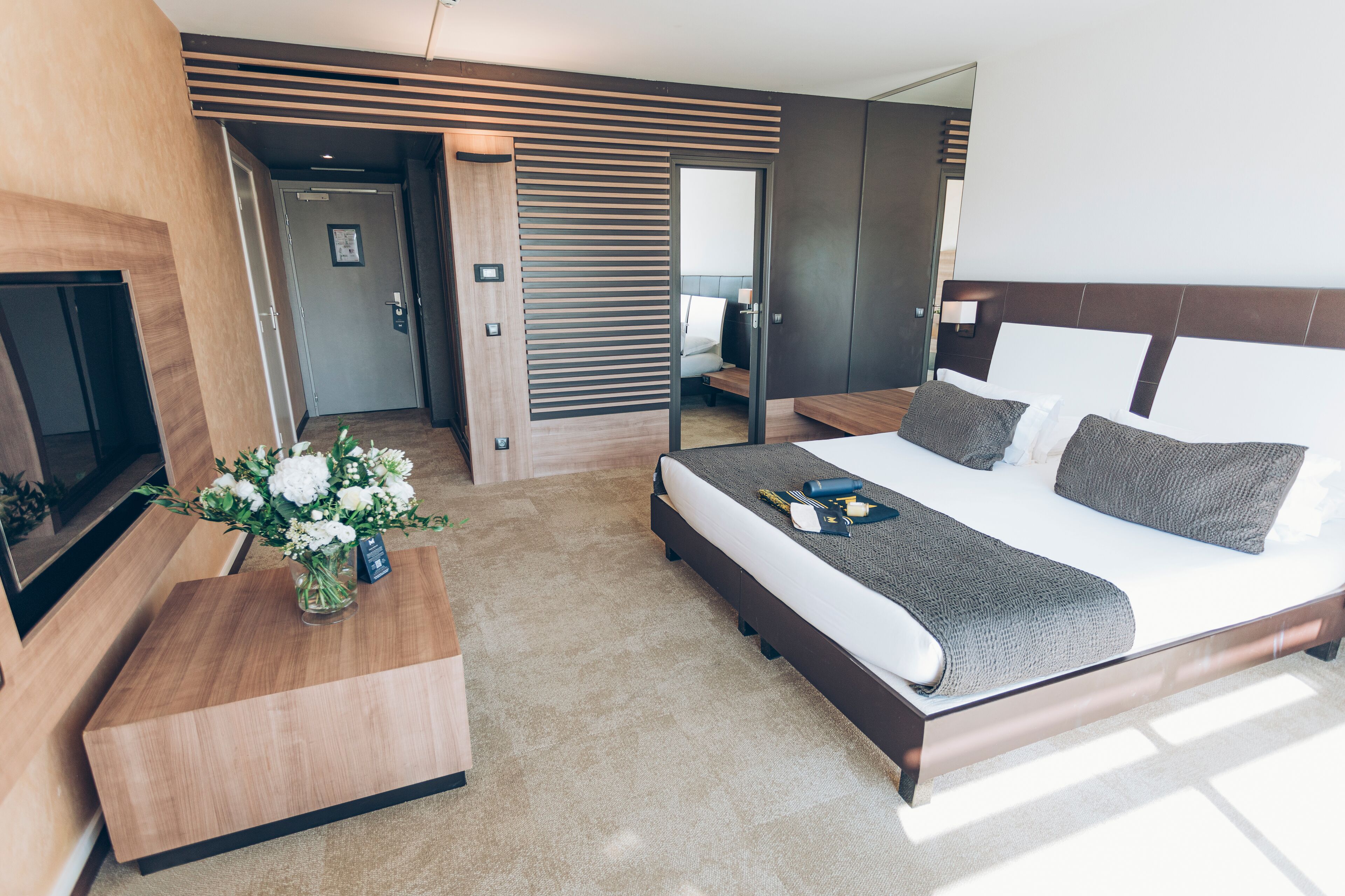 A modern hotel room with a large bed, a wooden nightstand, a TV, and a view of the city.