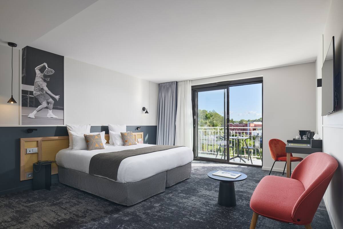 A modern hotel room with a large bed, a balcony with outdoor seating, and a flat-screen TV mounted on the wall.