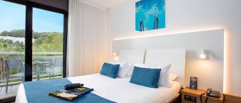 Habitación de hotel con una cama grande, vistas a la vegetación por la ventana, una pequeña mesa con sillas y una mesita de noche con un teléfono.