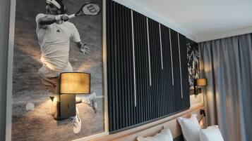 A modern hotel room with a large wall mural of a tennis player, a bed with white linens, a wooden headboard, and a black acoustic panel wall.
