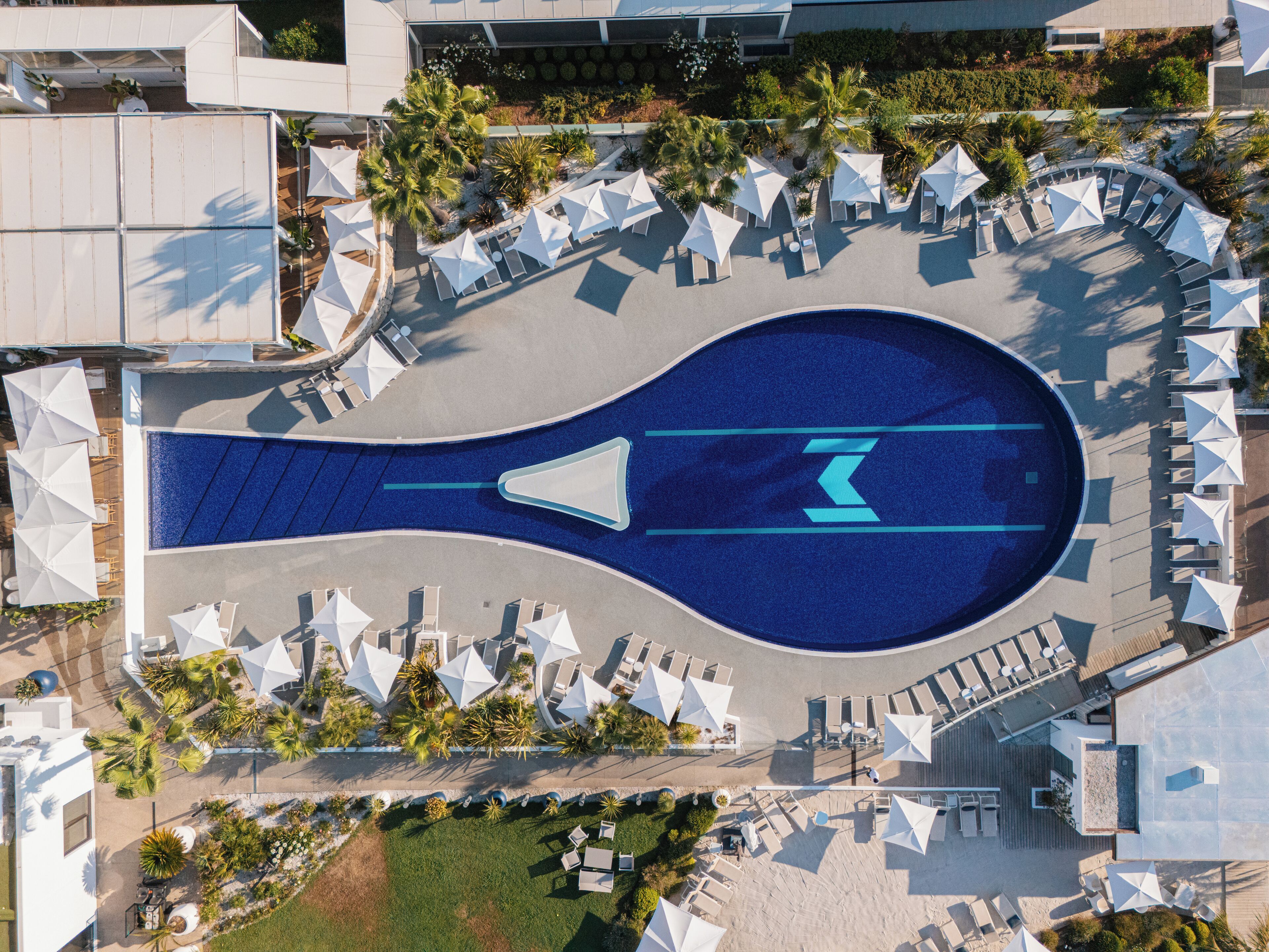 Luchtbeeld van een resort met een centraal zwembad, omringd door ligbedden en parasols, en diverse begroeiing.