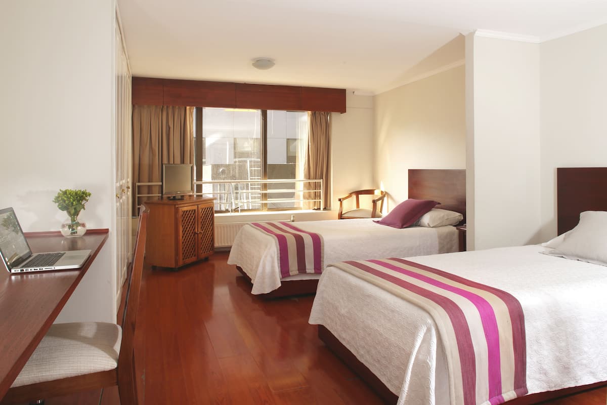 A hotel room with two beds, a desk with a laptop, a small table with a plant, and a view of the city through large windows.