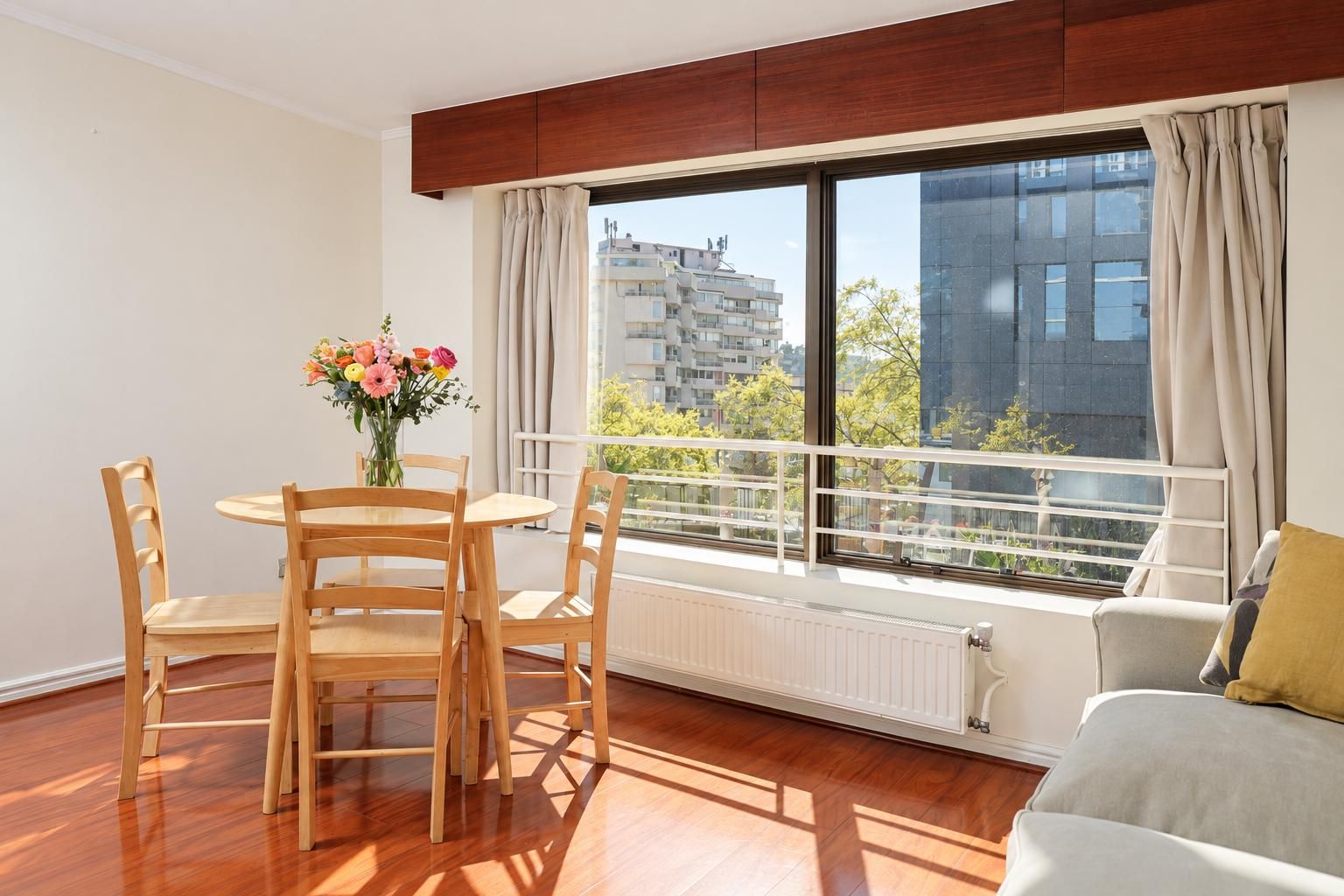 A modern apartment with a dining area, a sofa, a large window with a view of buildings, and a radiator.