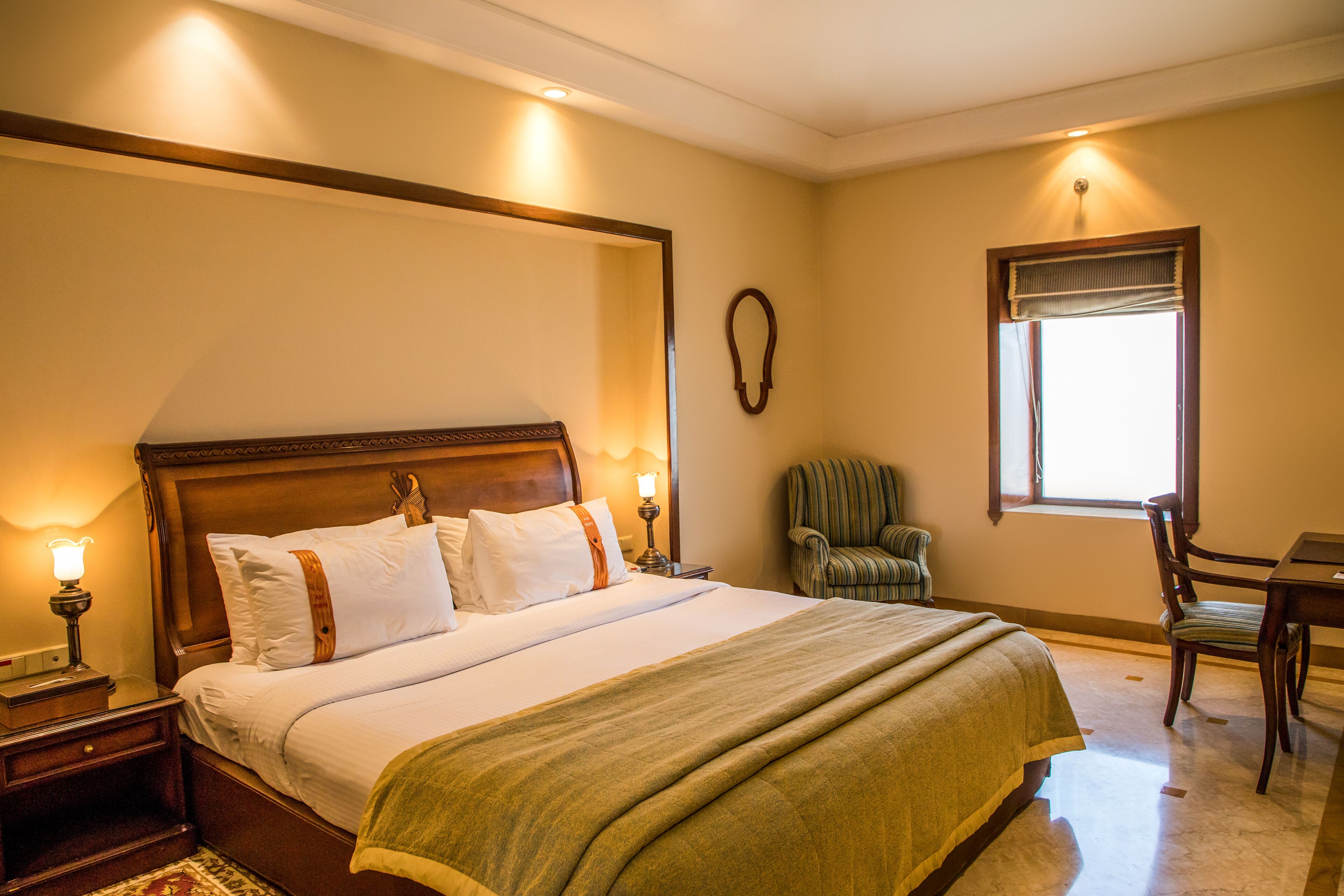 Una cama bien hecha con cabecera de madera, una mesita de noche con lámpara, una silla y una ventana con cortinas.