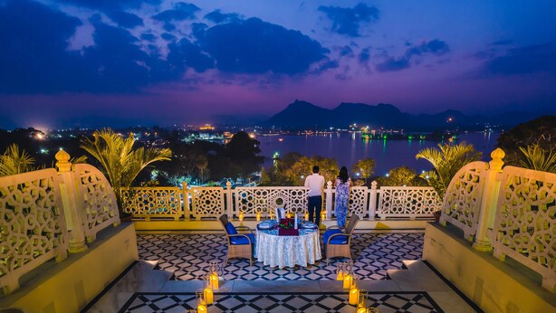 Une terrasse avec une table dressée pour deux personnes, offrant une vue sur un lac et le paysage urbain illuminé la nuit.