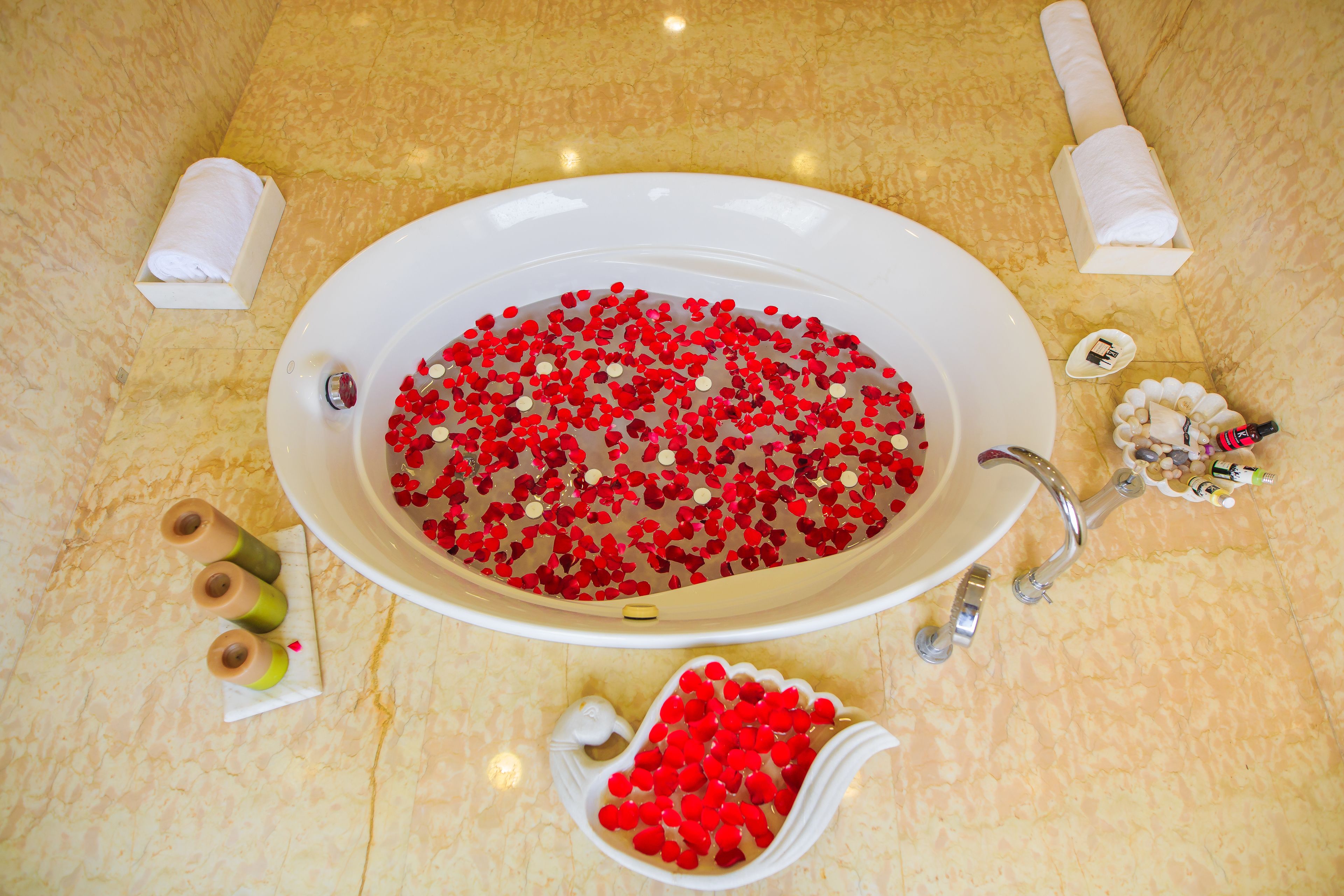 Une baignoire remplie de bombes de bain rouges en forme de cœur, entourée de serviettes blanches et de produits de bain, le tout posé sur un sol en marbre.