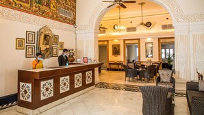 Un hall d’hôtel comprenant un comptoir d’accueil, des décorations murales élaborées, un lustre et un espace repas avec des meubles en osier.